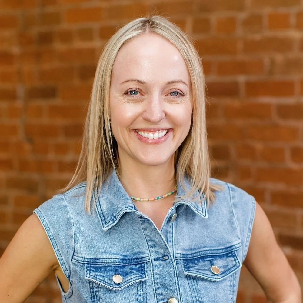 A smiling woman with shoulder-length blonde hair, wearing a sleeveless denim vest and a colorful beaded necklace, standing in front of a brick wall.