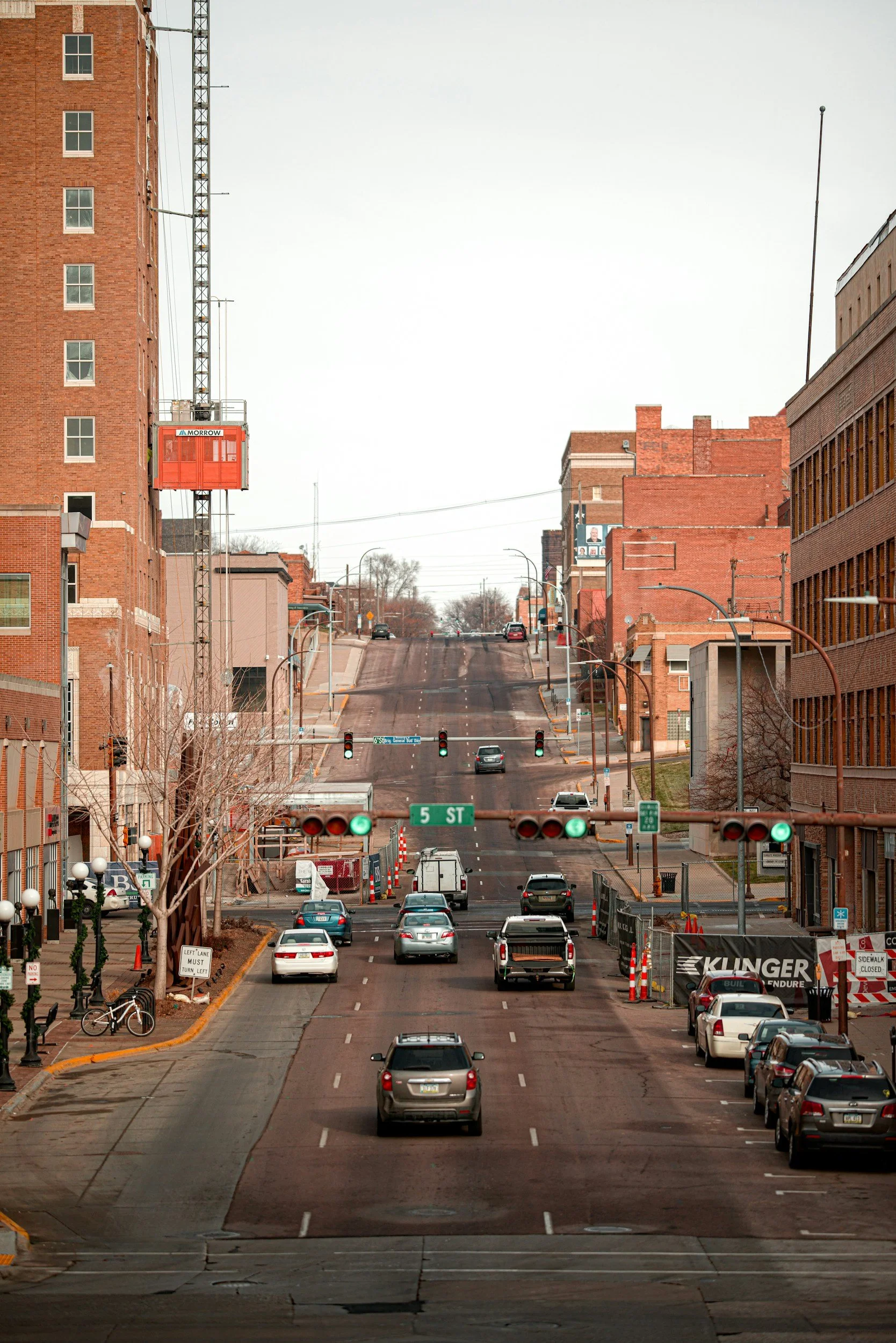 City street with cars waiting at a traffic light on 5th Street, surrounded by brick buildings, streetlights, and a construction site on the right.