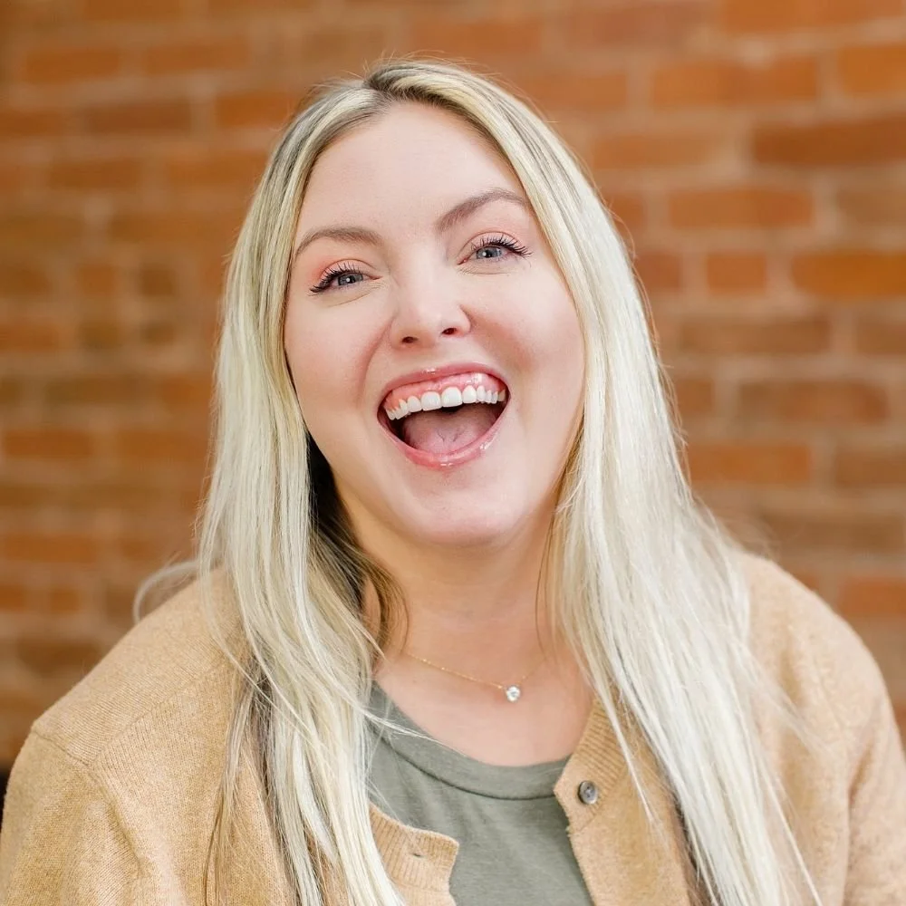 Close-up portrait of a young woman with long blonde hair, smiling with an open mouth, against a brick wall background.