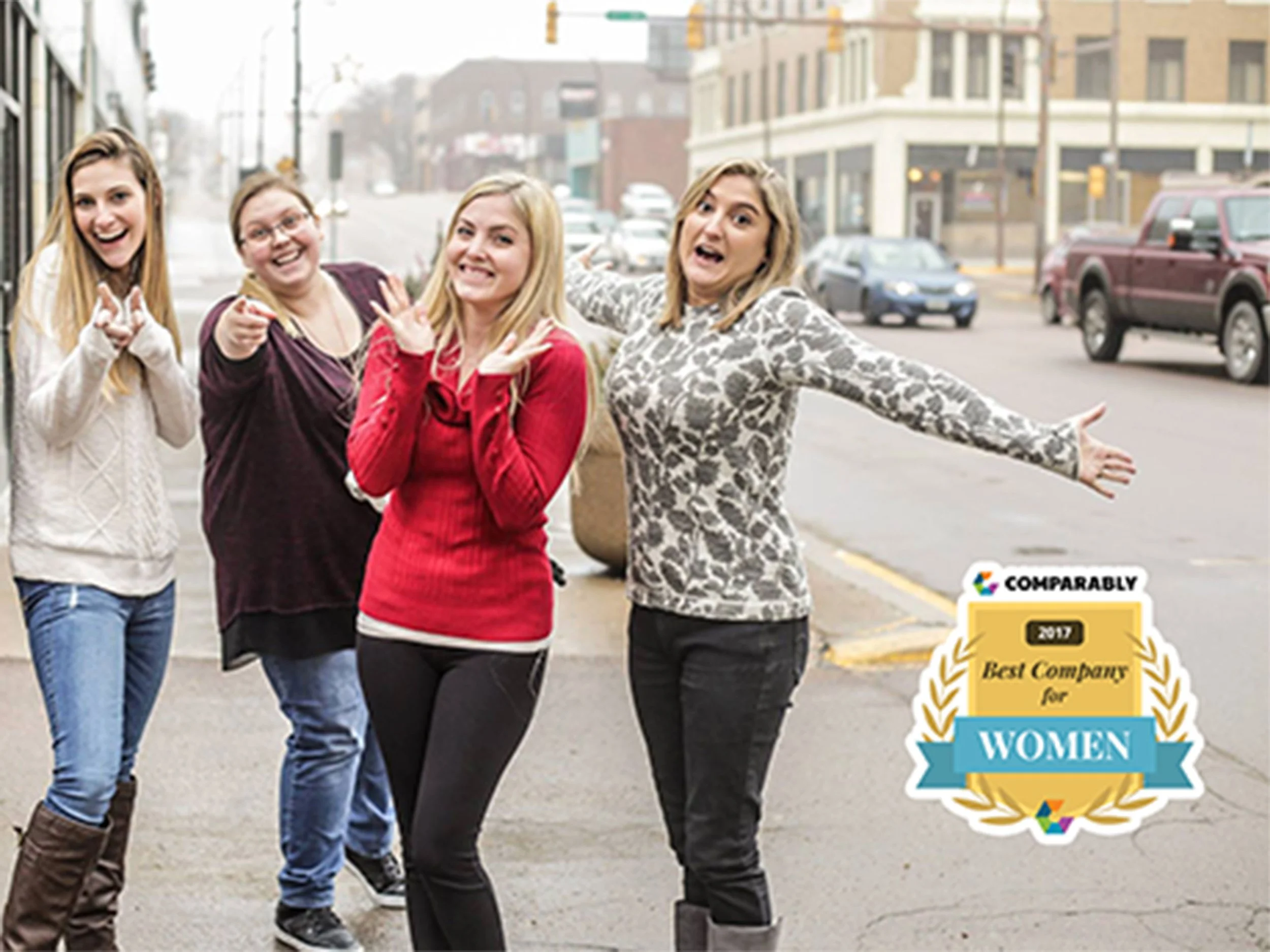 Four women standing on a city sidewalk, smiling and making playful gestures. One woman has her arms outstretched. The image has a badge that says "2017 Best Company for Women."