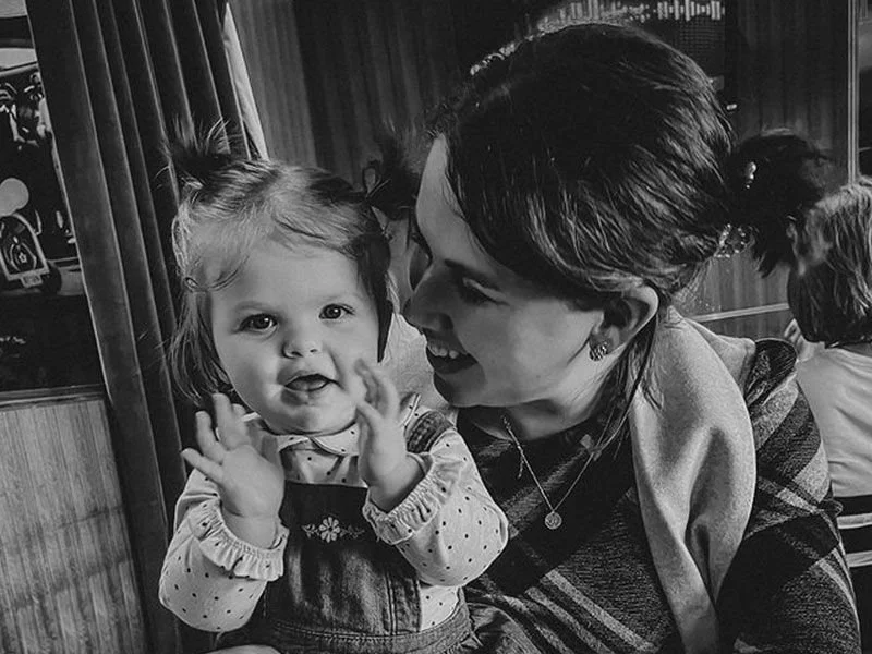 A woman smiling and looking at a young girl with her hands raised in a playful gesture, in a cozy interior setting.
