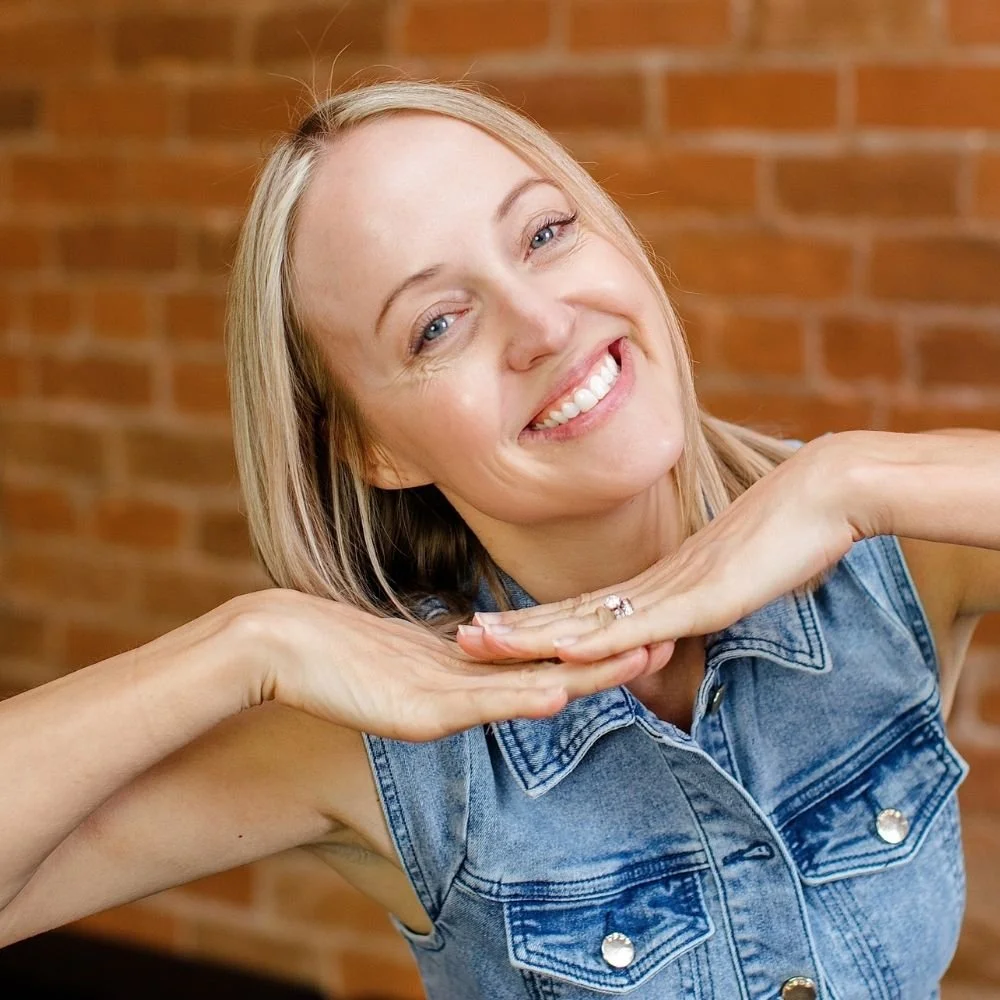A woman smiling and posing with her hands under her chin in front of a brick wall.
