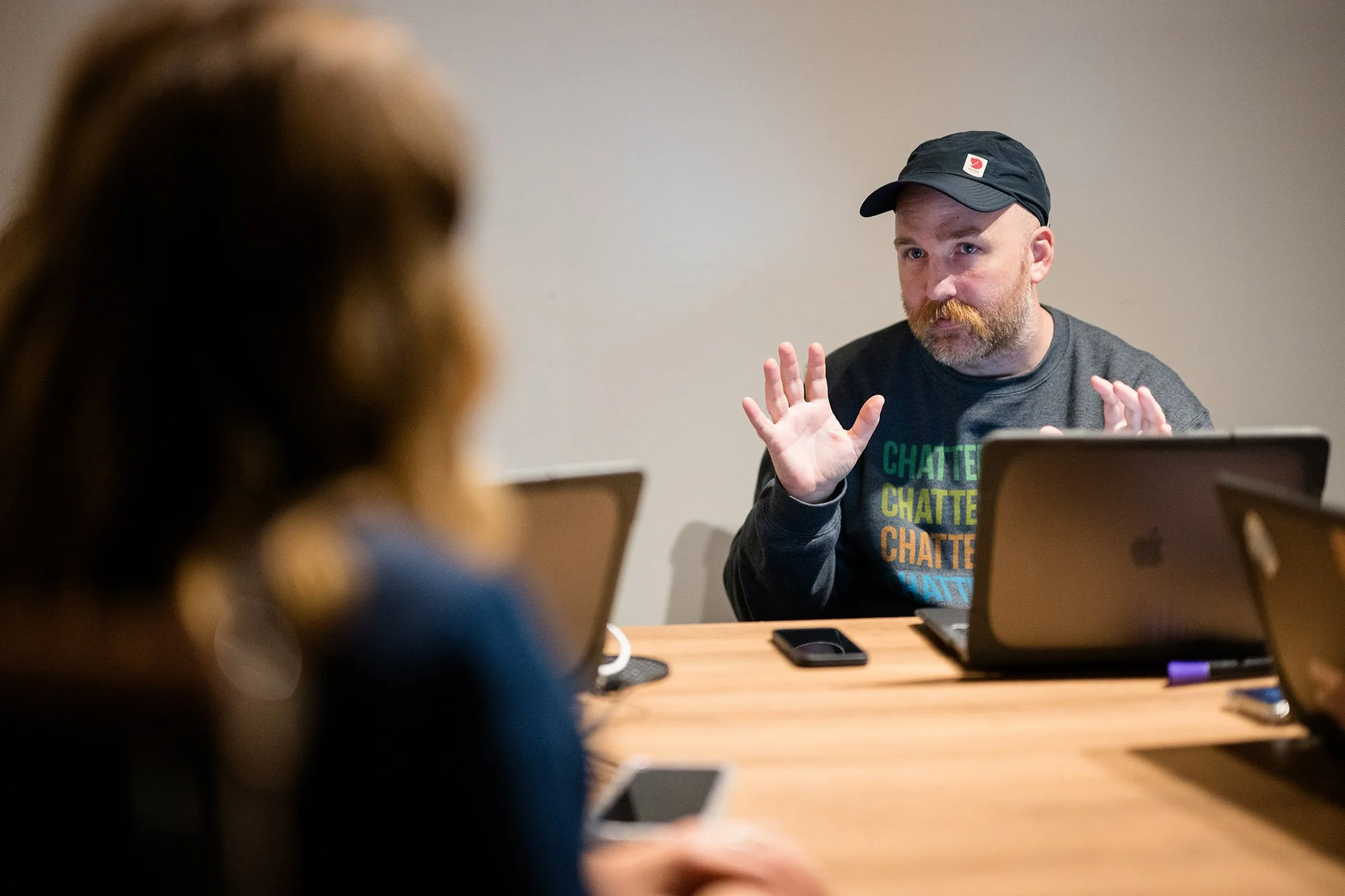 A man with a beard, wearing a gray shirt with colorful text and a black cap, is gesturing with his hands while sitting at a table with laptops, in a meeting room with a plain wall.
