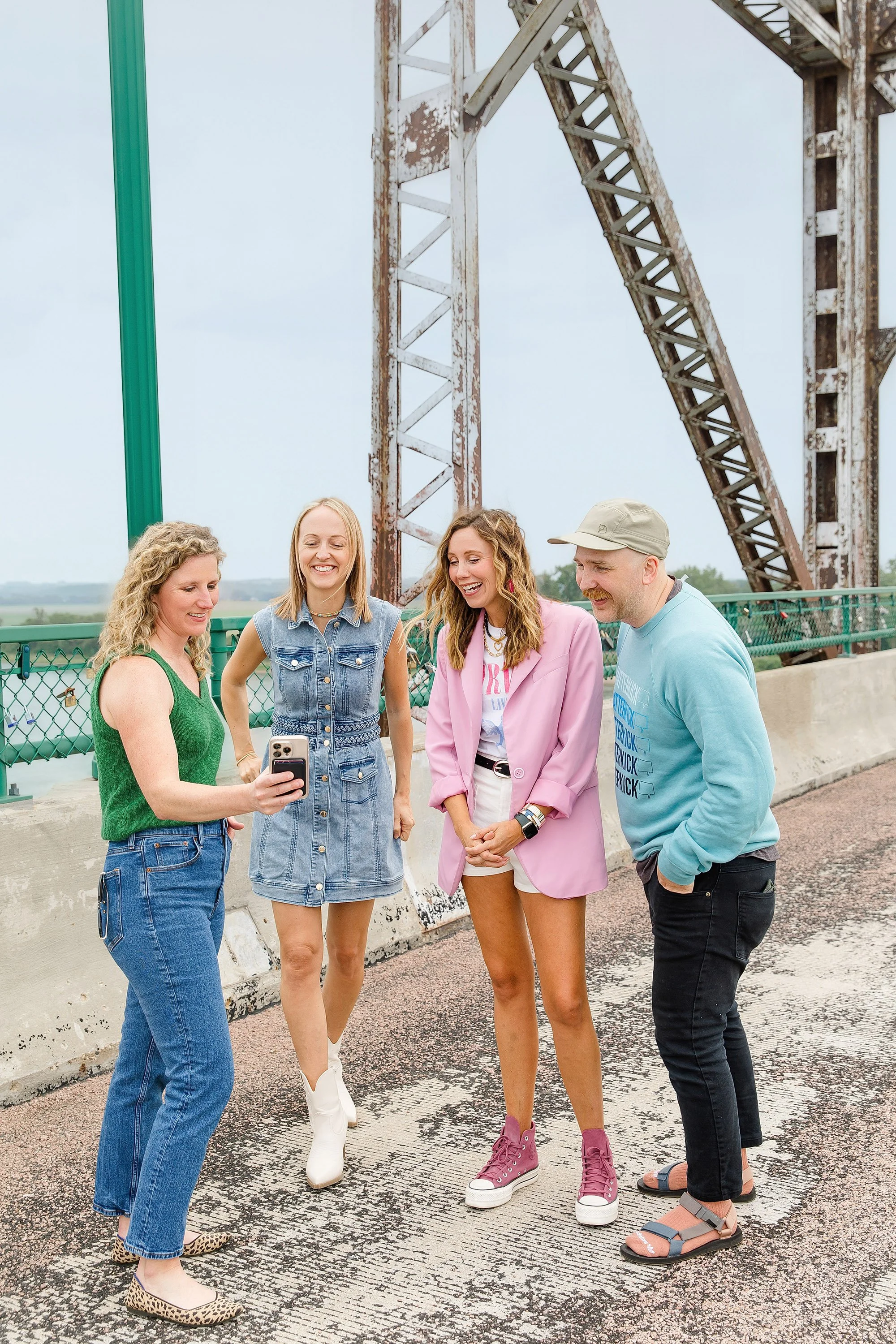 Four young adults standing on an outdoor bridge, smiling and looking at a smartphone, with a rusted metal structure in the background.