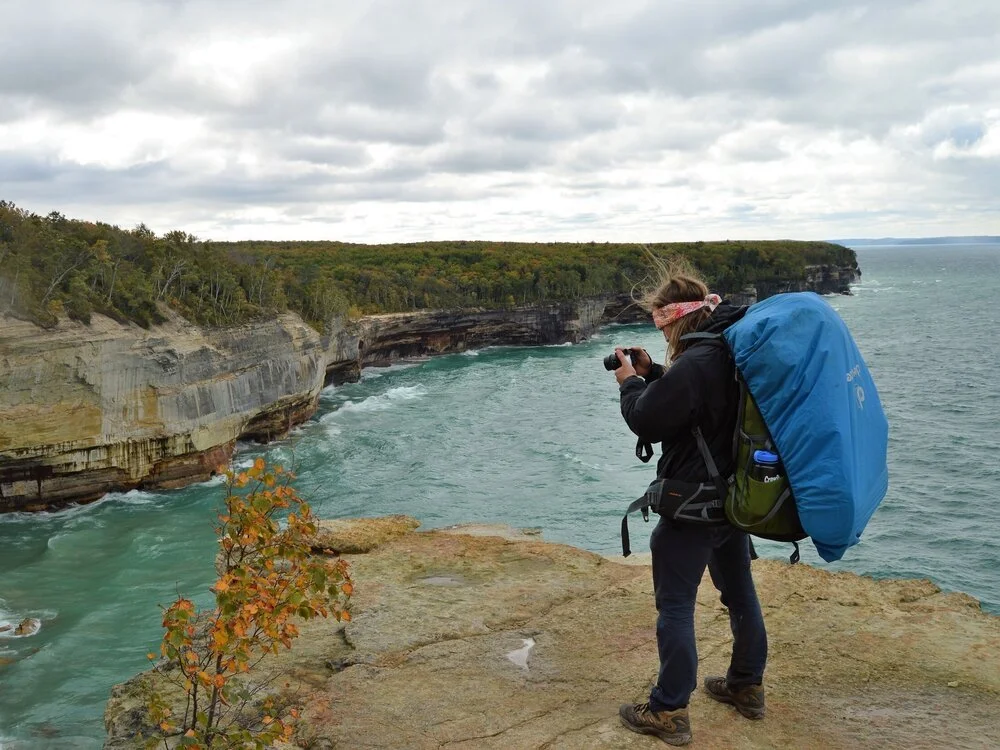 Person with a blue backpack taking photos near a rocky cliff overlooking a turquoise river and forested landscape.
