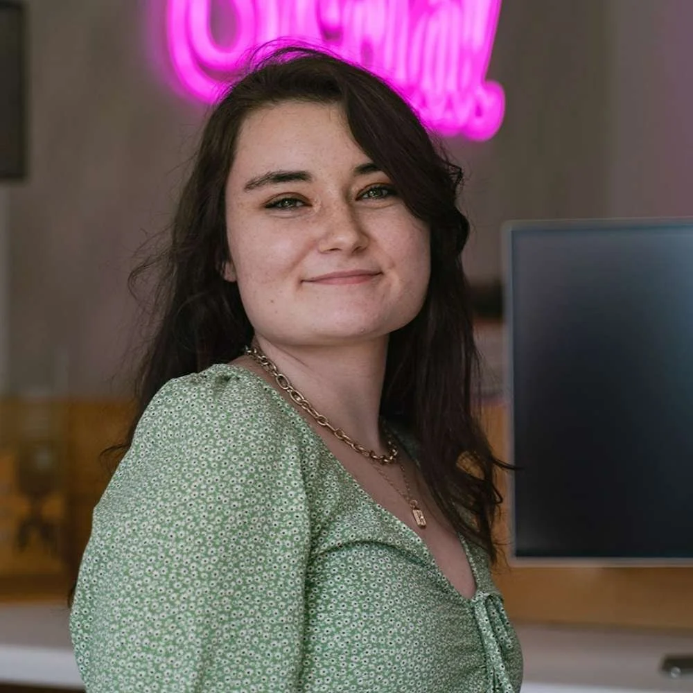 A woman with dark hair, wearing a green patterned top and gold chains, smiling in a cafe with a pink neon sign in the background.