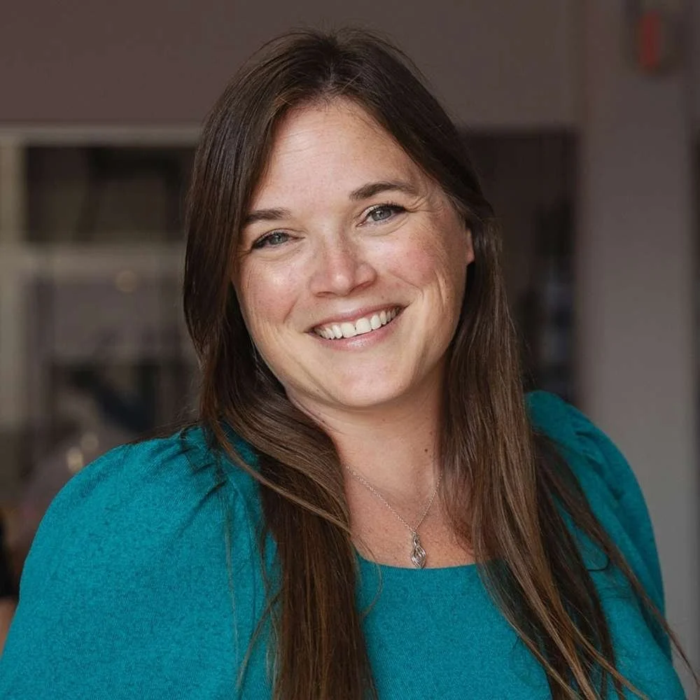 A woman with long brown hair smiling, wearing a teal top and a silver necklace, in an indoor setting.