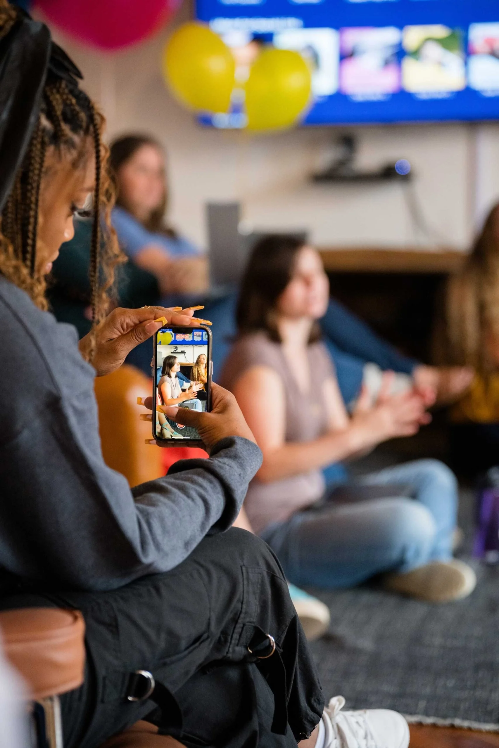A woman with braids taking a photo with her phone of people sitting on a bench at a social event.