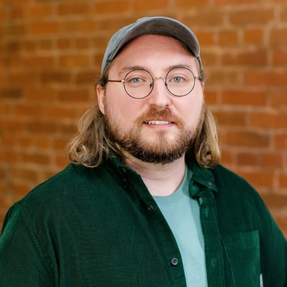 A man with long hair, wearing glasses, a gray cap, a green shirt, and a light-colored t-shirt standing in front of a brick wall.
