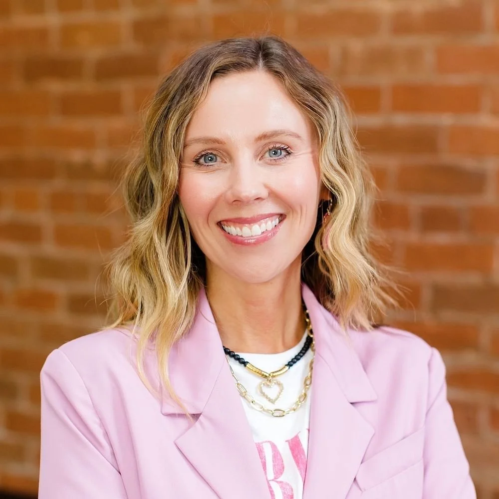 A woman with wavy blonde hair smiling, wearing a pink blazer, in front of a brick wall background.