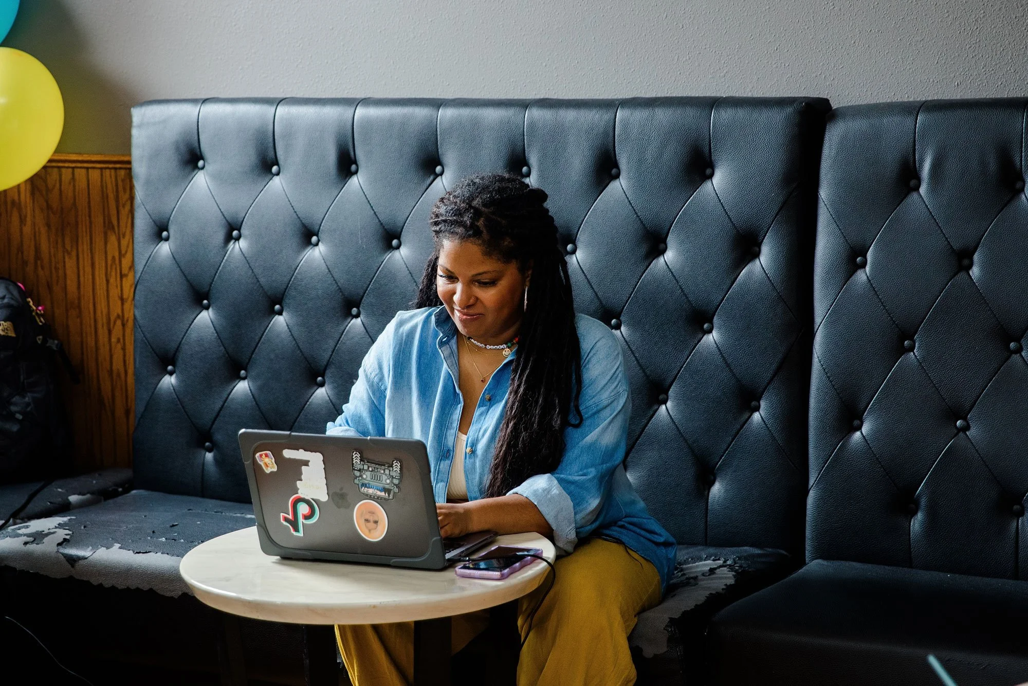 Woman sitting on a black tufted booth using a laptop at a round white table.