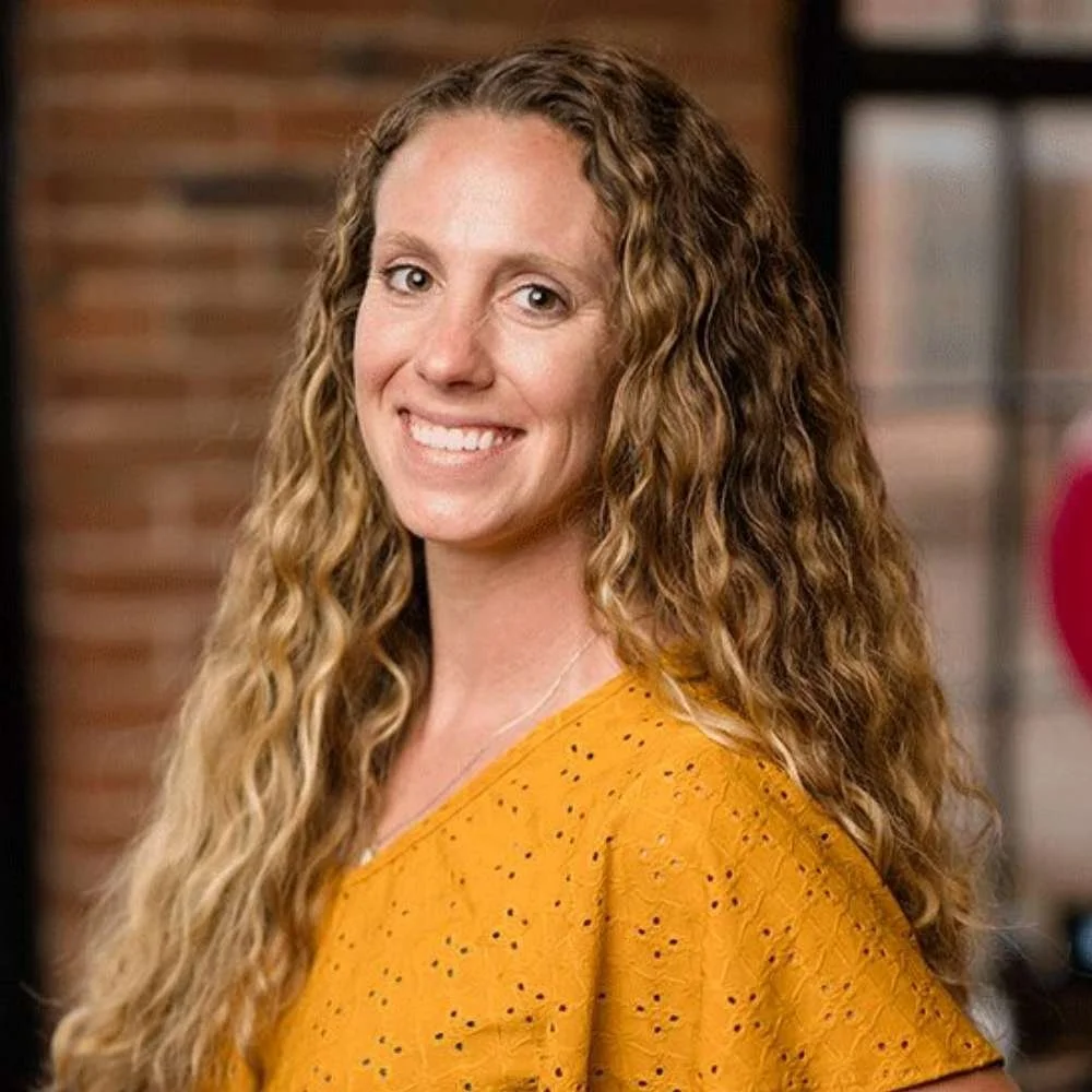 A woman with long, curly blonde hair smiling in an indoor setting, wearing a mustard yellow top with small holes.