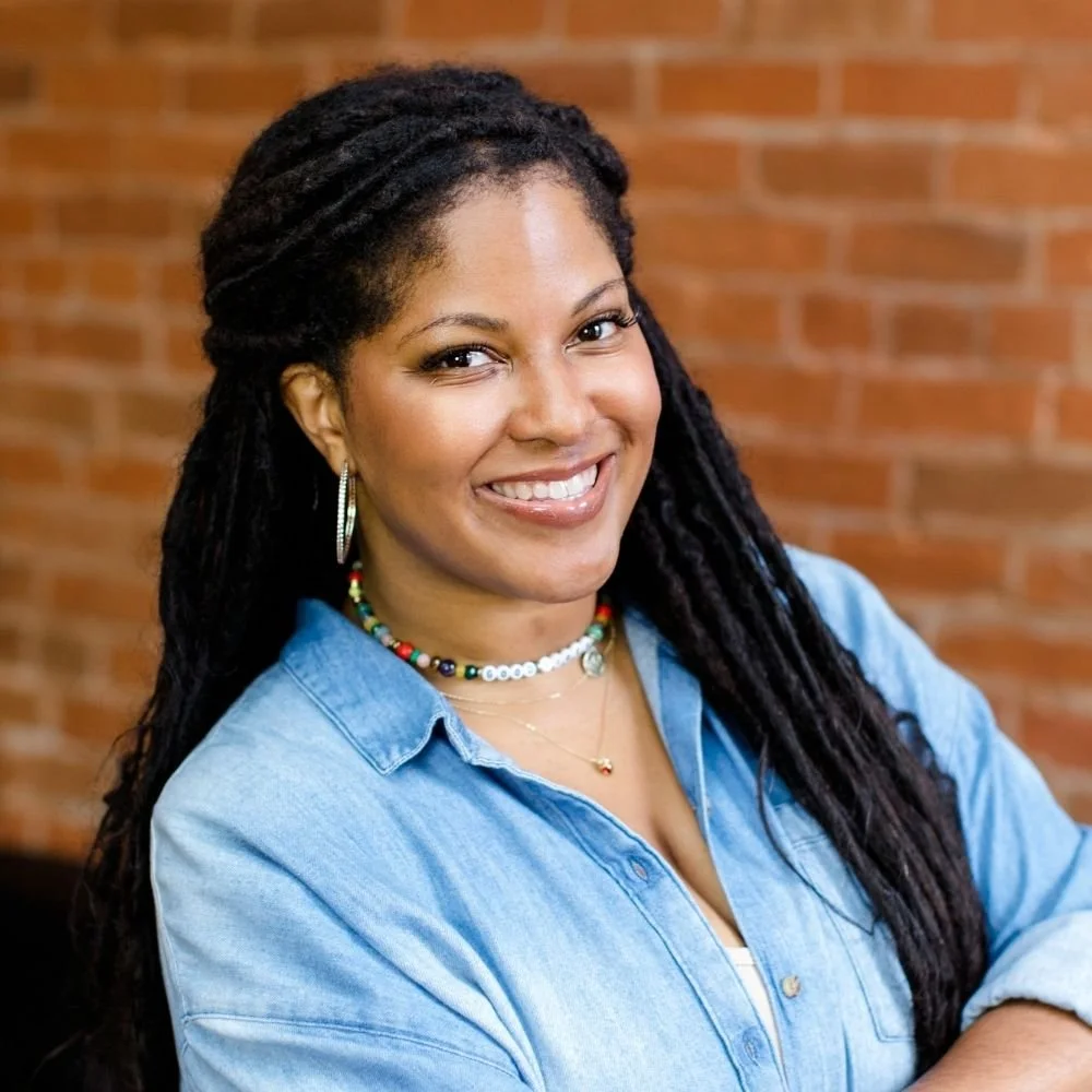 A woman with long curly hair, smiling, wearing a denim shirt, colorful beaded necklaces, hoop earrings, and a white top, posing against a brick wall background.
