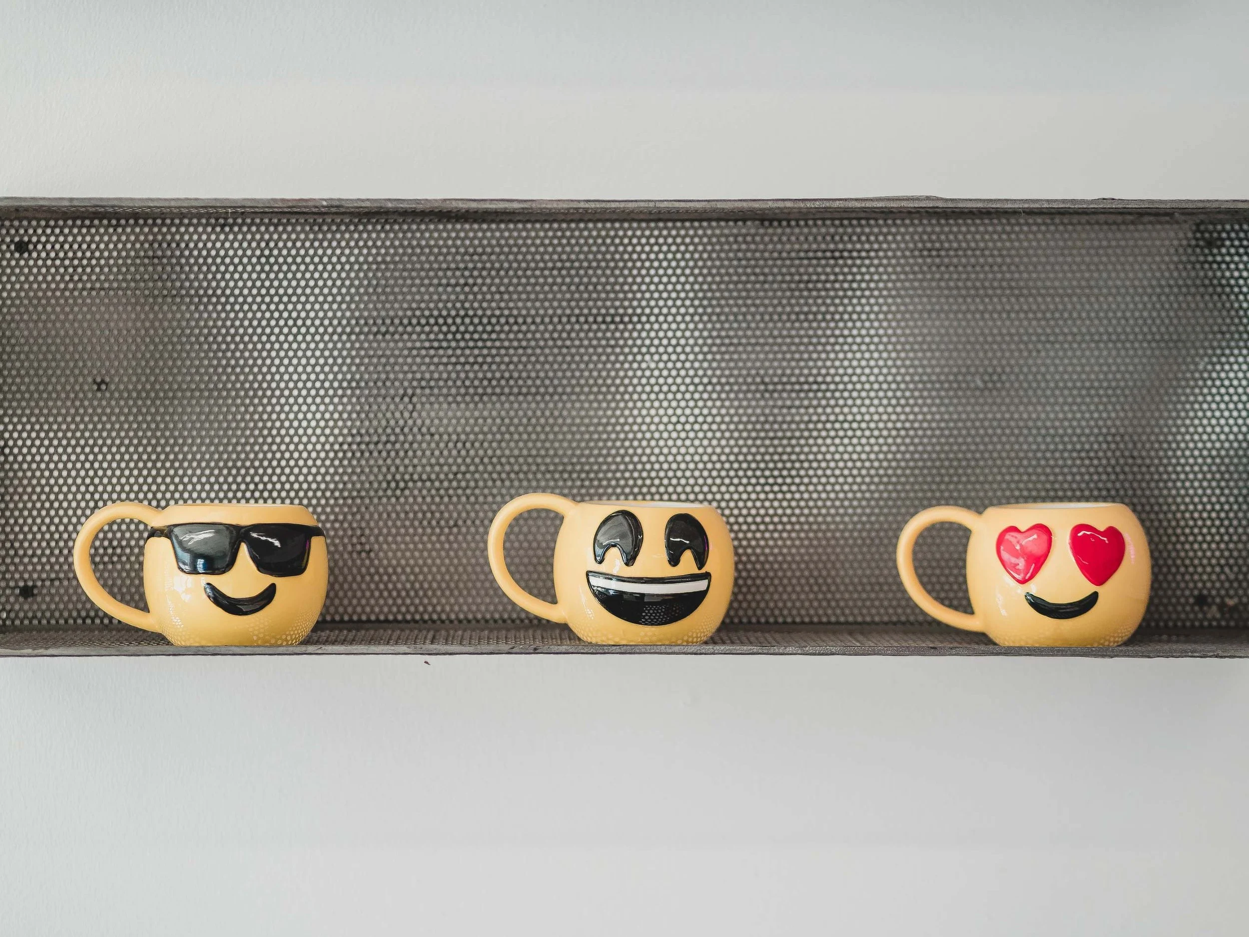Three yellow coffee mugs with emoji faces, displayed on a shelf with a metallic backing; the first mug has sunglasses, the second mug has a big smile with closed eyes, and the third mug has heart eyes.
