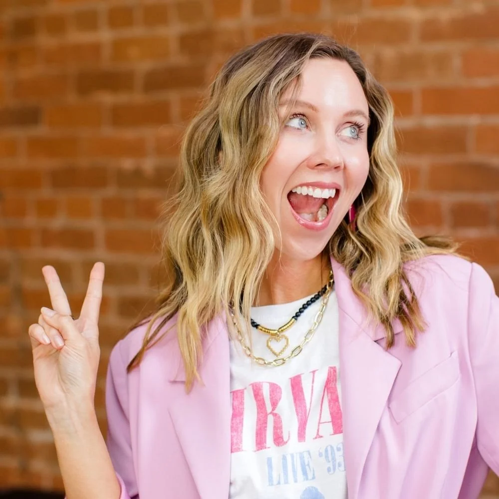 Woman with wavy blonde hair wearing a pink blazer, white Nirvana t-shirt, layered necklaces, making a peace sign gesture, smiling with her mouth open, standing in front of a brick wall.
