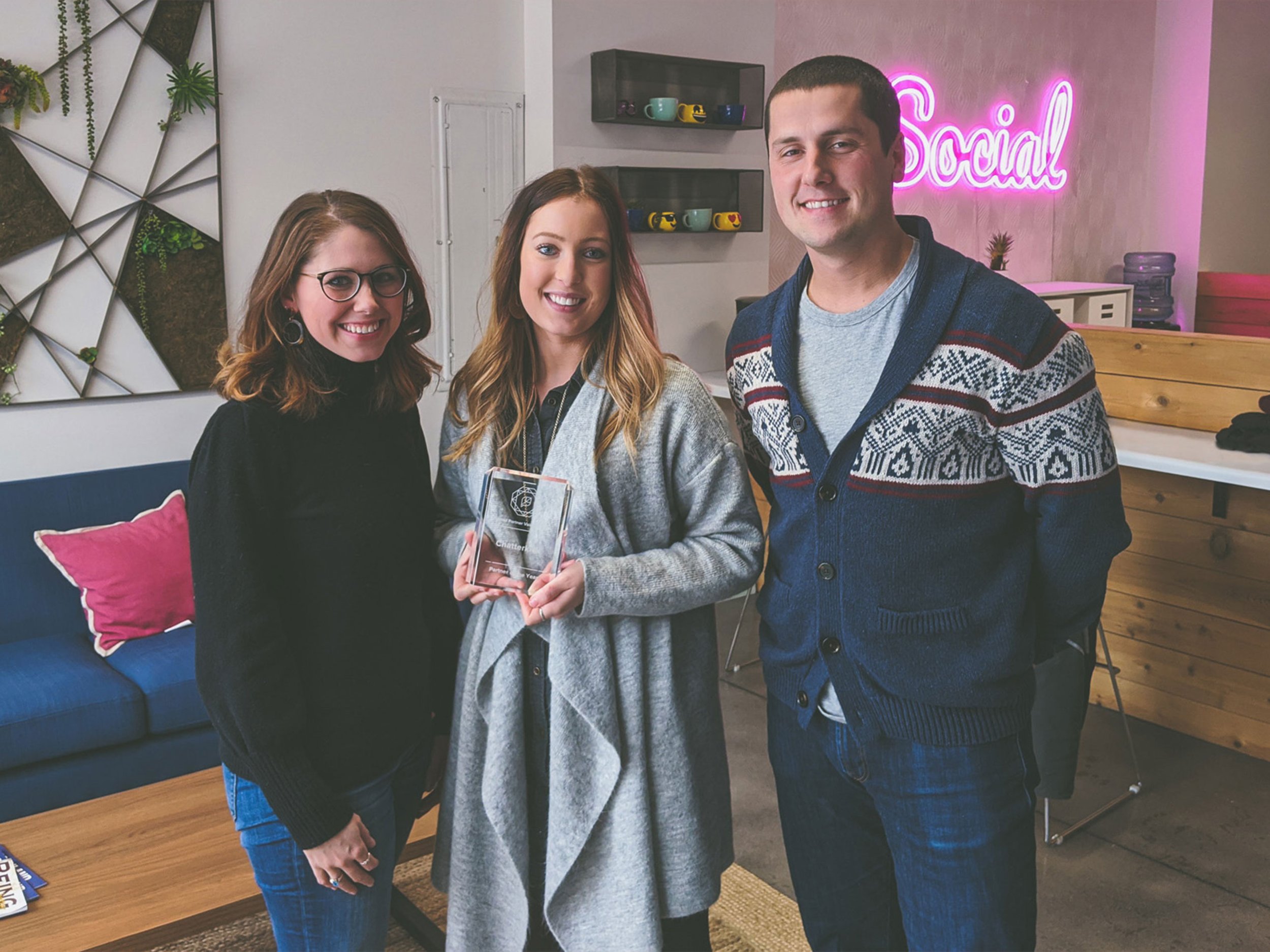Three smiling people inside a modern office space, one woman holding a clear rectangular award, pink and purple neon 'Social' sign in the background, and a decorative wall with green plants.