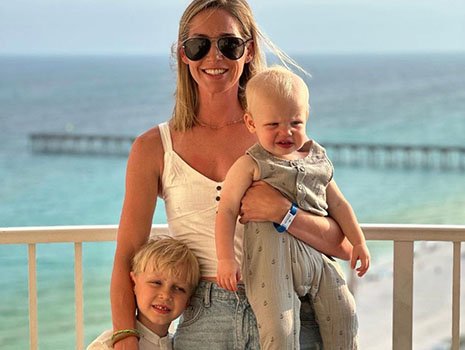 A woman with sunglasses holding two young children on a beachside balcony with the ocean and pier in the background.