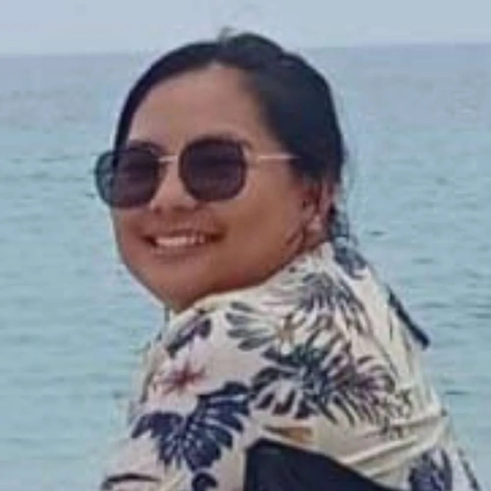 A woman with dark hair and sunglasses smiling at the camera, wearing a printed shirt, with a body of water in the background.