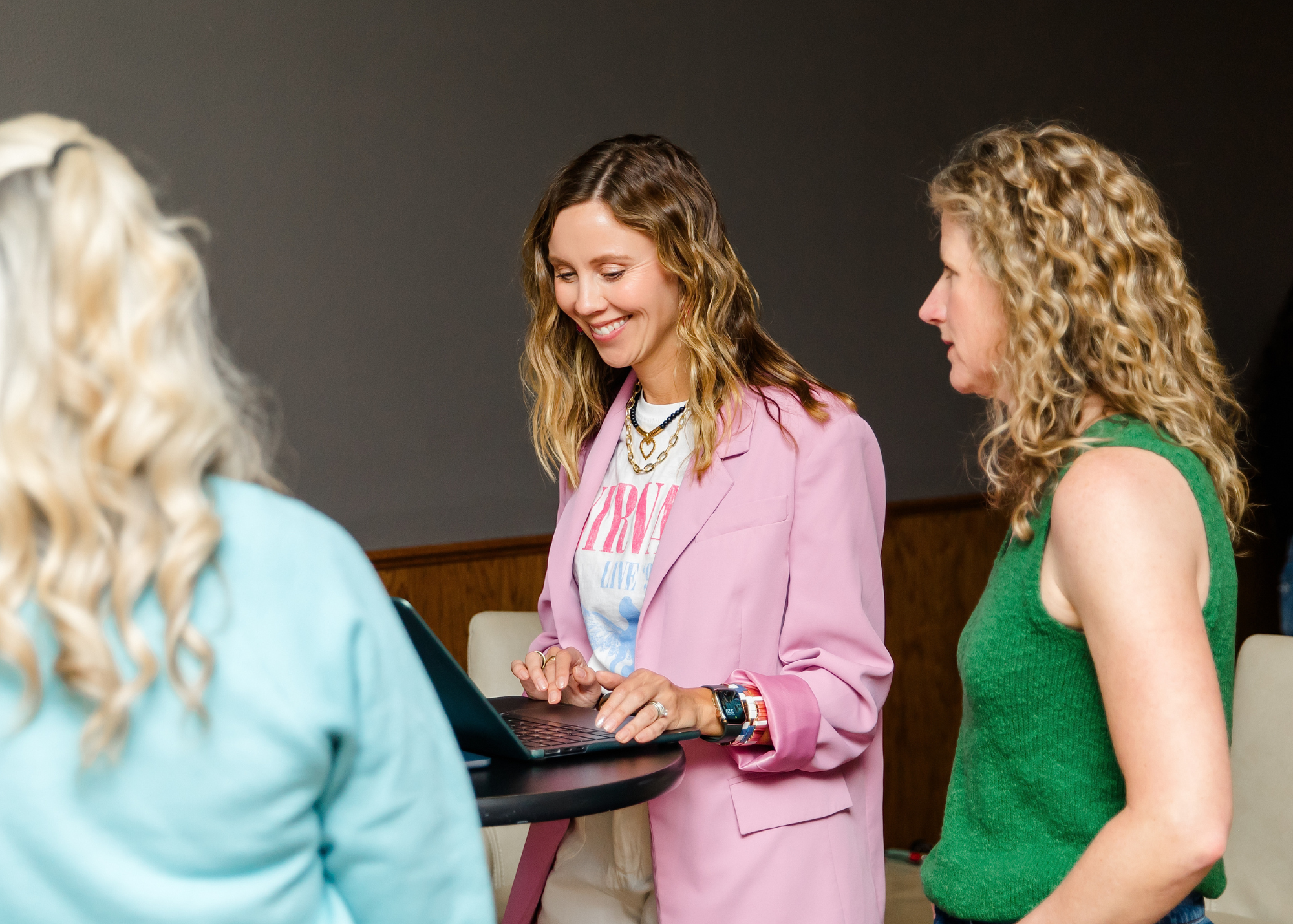 Three women having a conversation indoors, one using a laptop, casual social setting