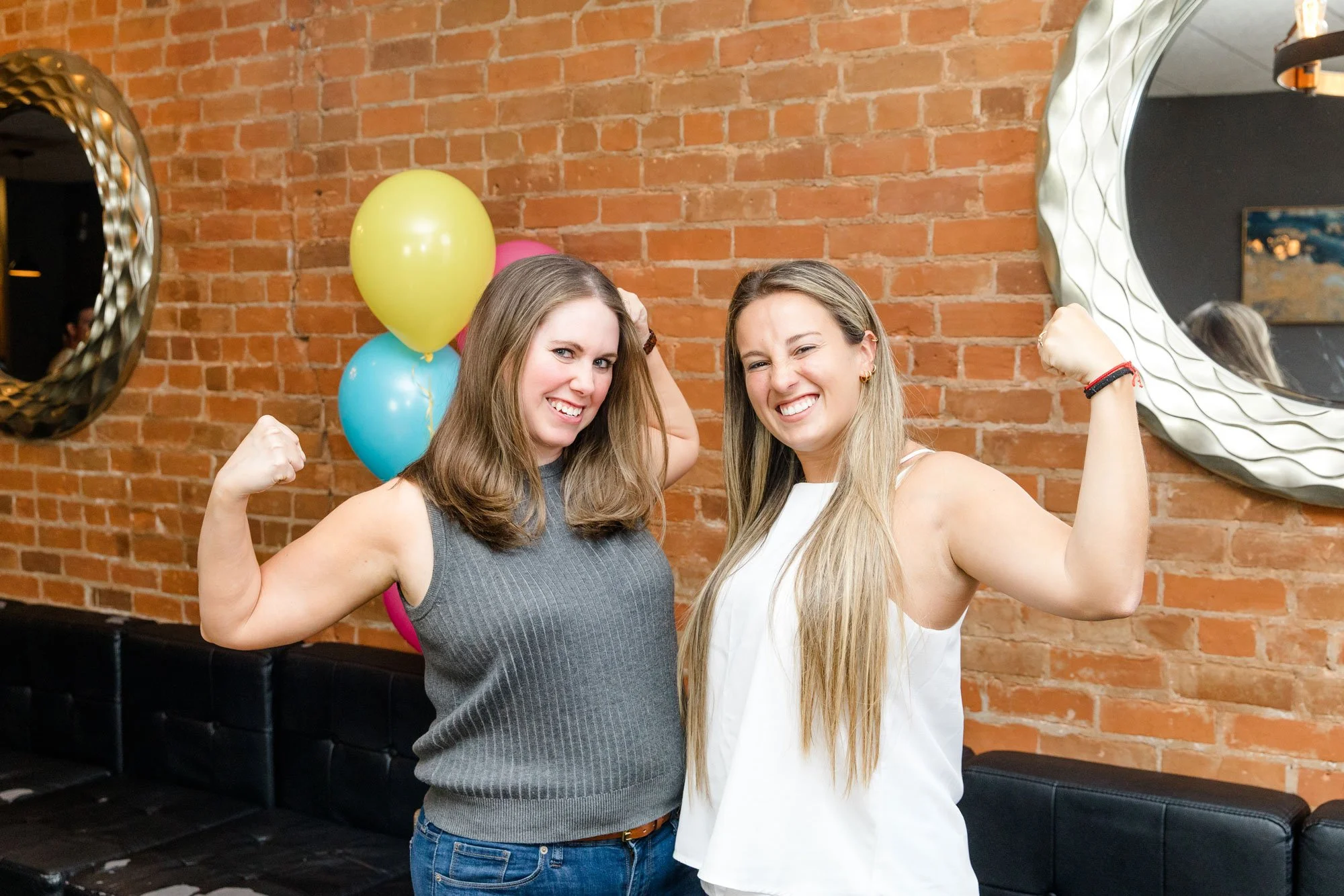 Two women flexing their biceps and smiling at a celebration with balloons in the background, against a brick wall.