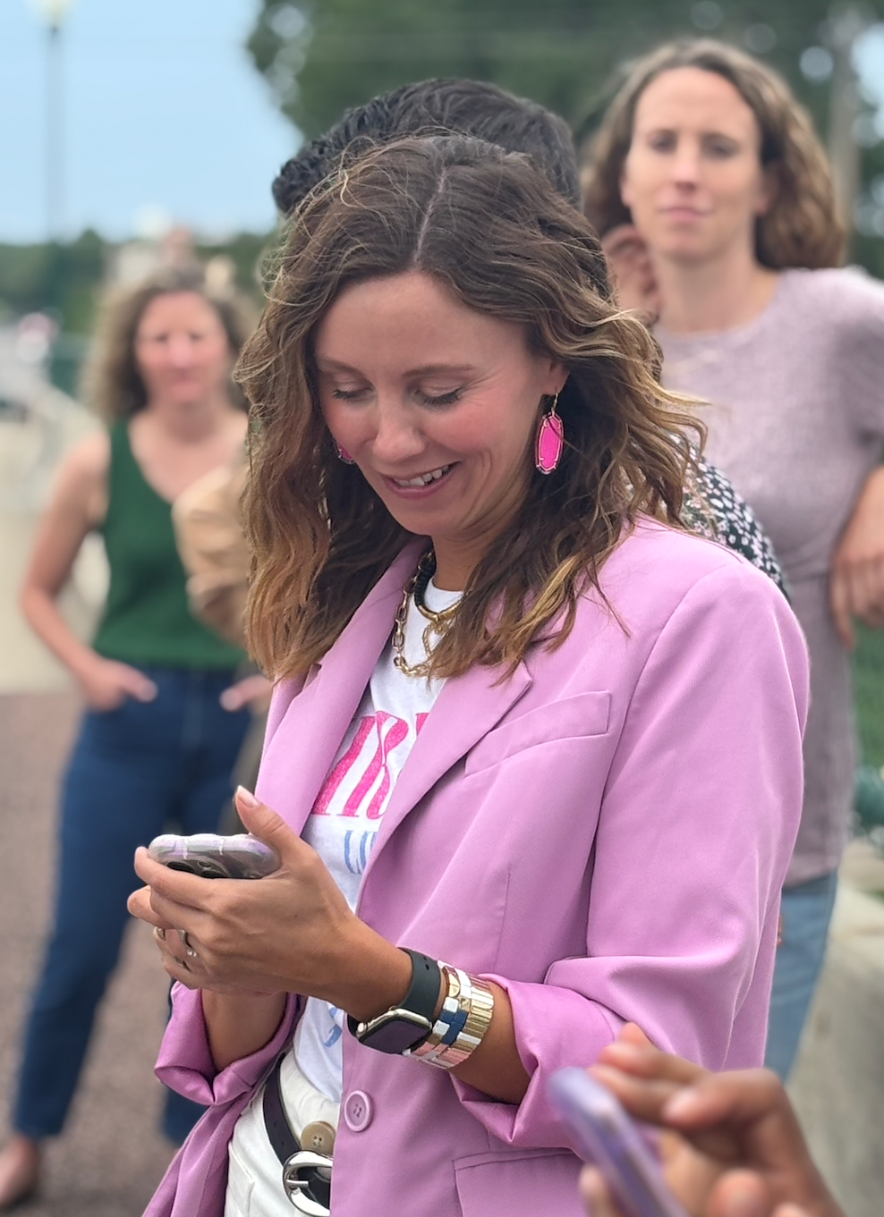 A woman with brown wavy hair wearing a pink blazer, pink earrings, white t shirt, and gold jewelry looking at her phone while smiling at an outdoor gathering with others in the background.