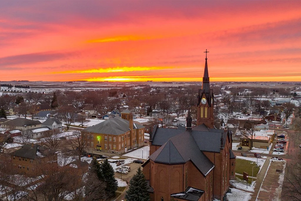 A small town at sunset with a colorful sky, featuring a prominent red brick church with a tall steeple and cross, surrounded by snow-covered buildings and streets.