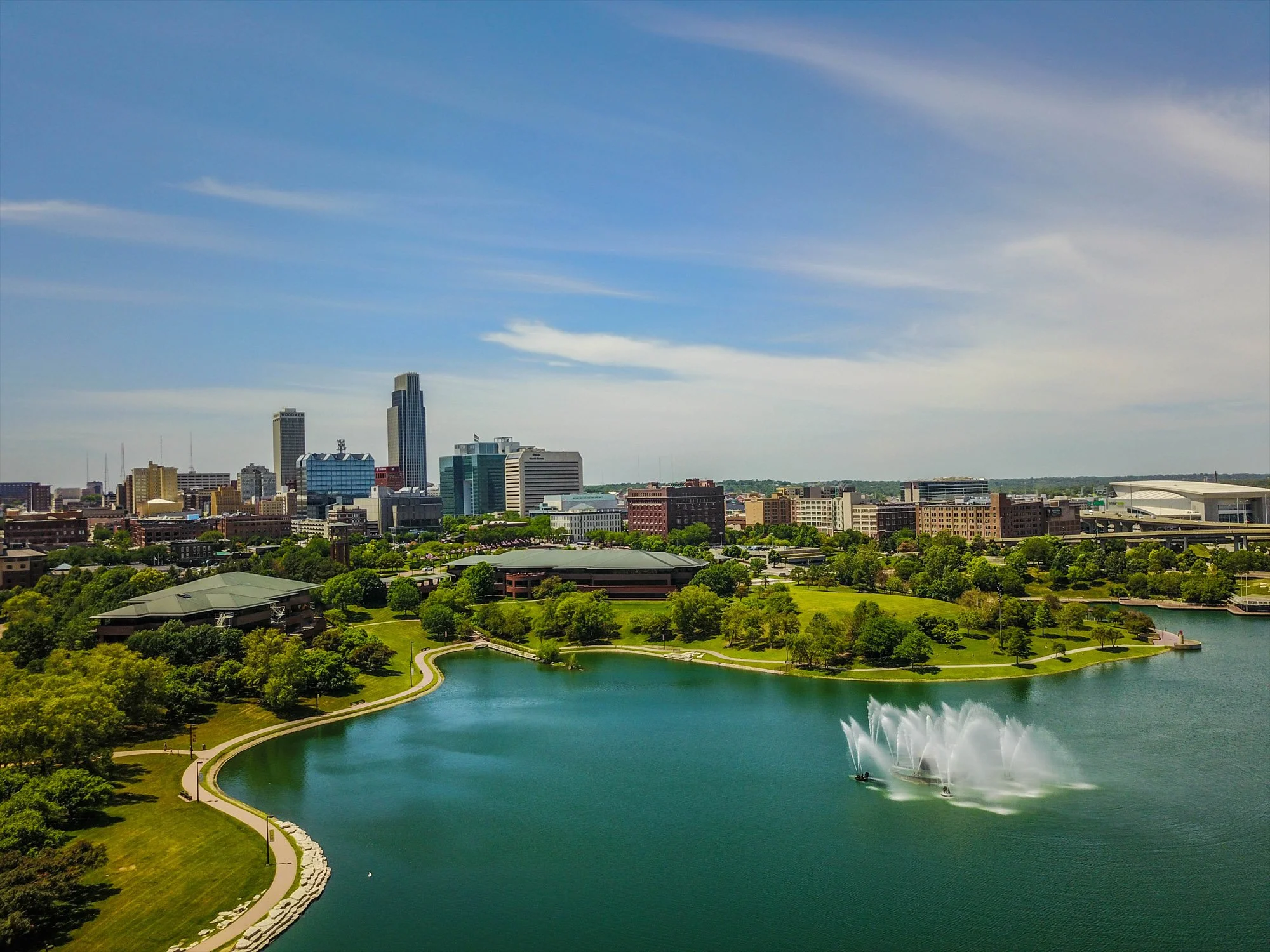City skyline with modern high-rise buildings, a large body of water with a fountain, green parks, and a clear blue sky.