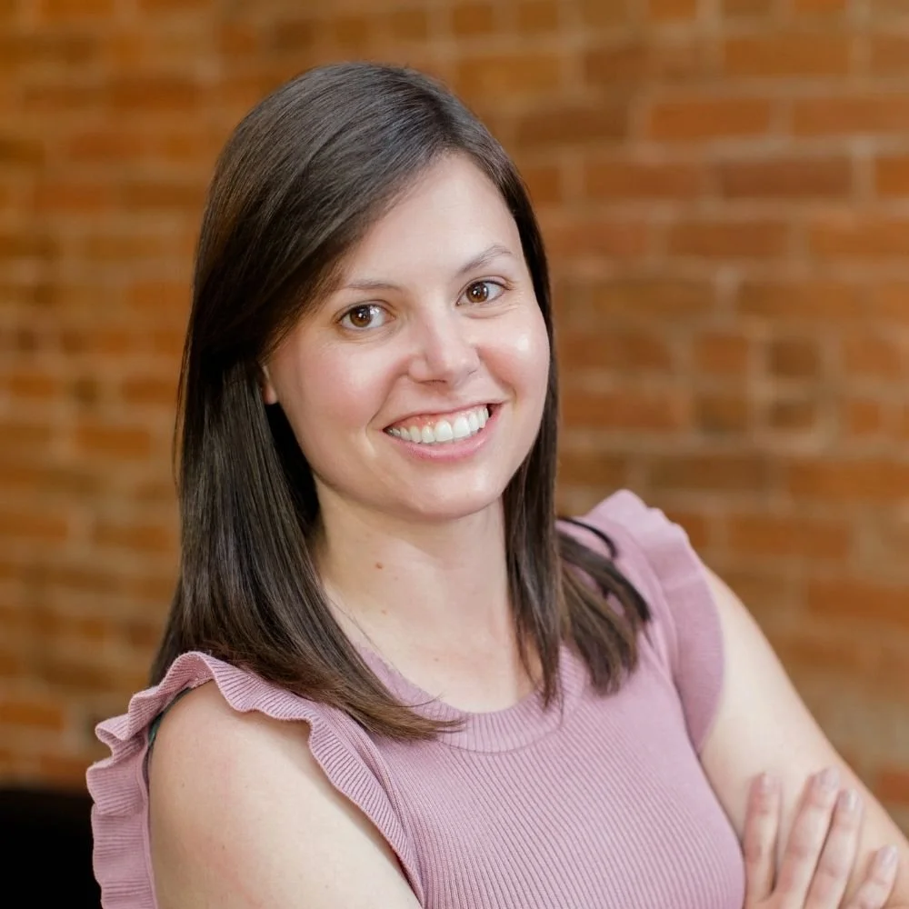 A smiling woman with brown hair wearing a pink sleeveless top with ruffled shoulder details, standing with crossed arms in front of a brick wall.