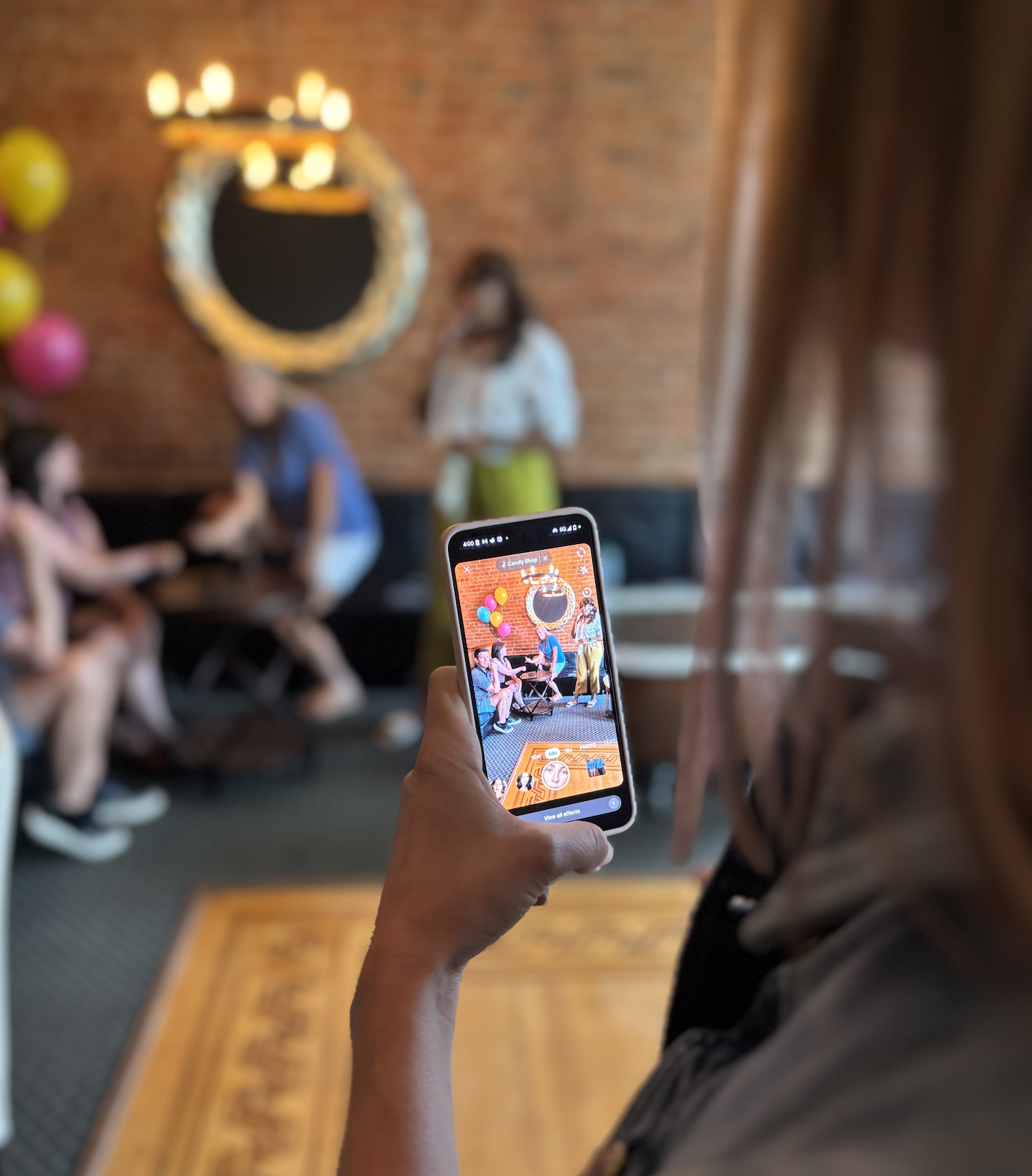 Person taking a photo of a group of people at a birthday party with balloons and a brick wall background.