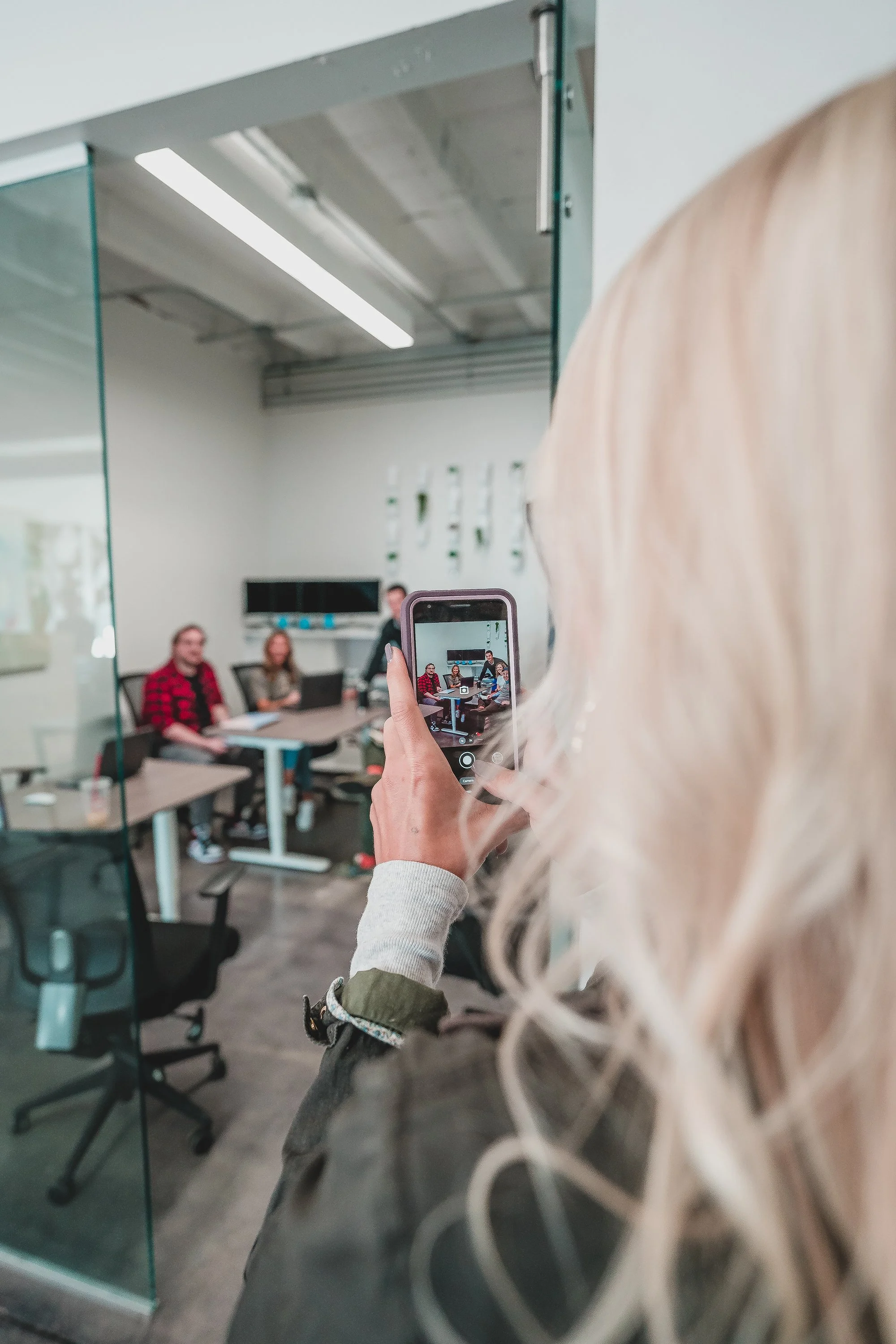 A woman with blonde hair taking a photo of a group of four people sitting around a conference table in a modern office conference room.