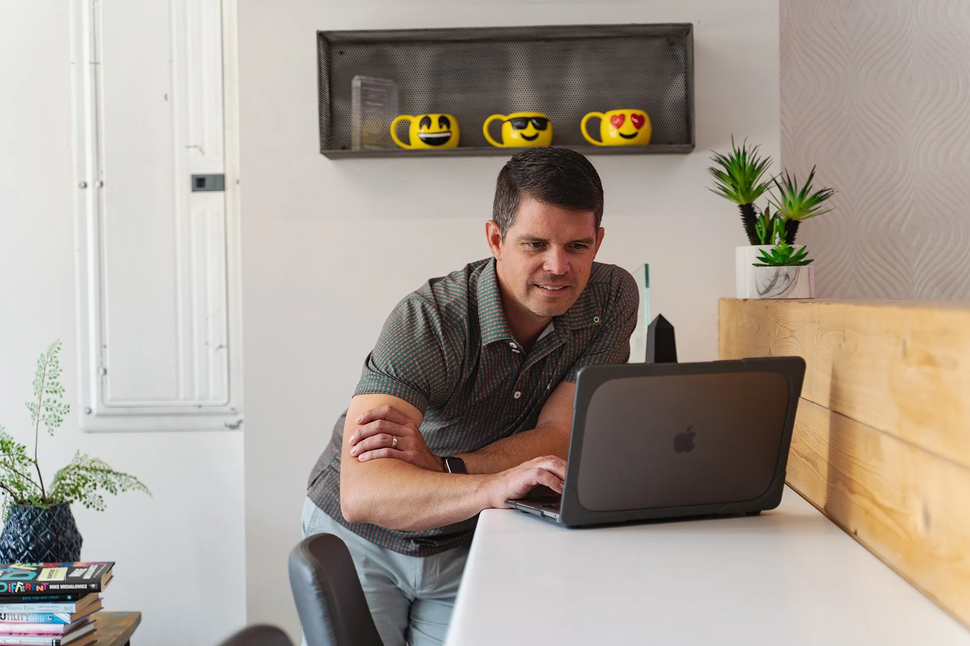 A man looking at a laptop on a white desk, leaning forward with his arms crossed, in a modern room with plants and a shelf with smiley mug decorations in the background.