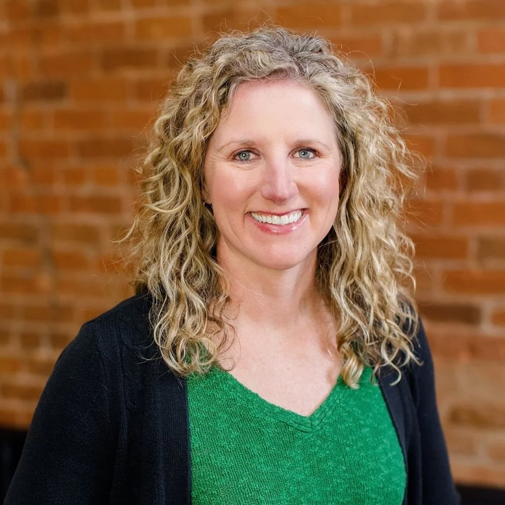 A woman with curly blonde hair wearing a green top and black cardigan, smiling in front of a brick wall.
