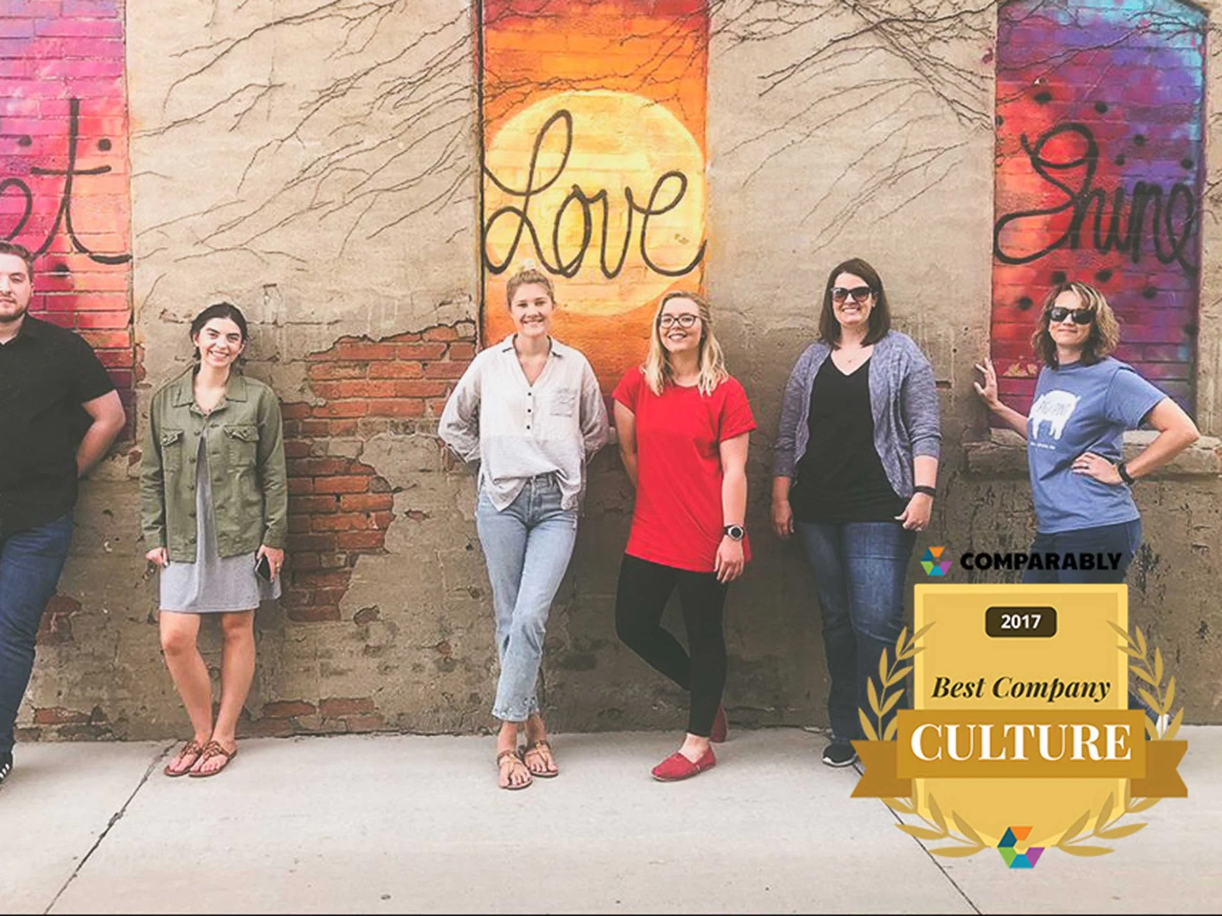 Six women stand in front of a brick wall with colorful painted panels. The panels have the words 'Love' and 'Share' painted on them. An award graphic overlay indicates the company was recognized in 2017 as the 'Best Company Culture' by Comparably.