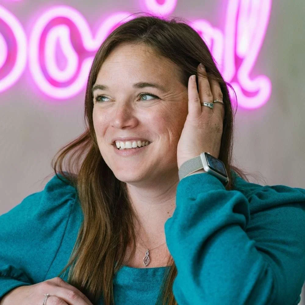 A woman with brown hair and blue eyes smiling, wearing a teal top and a smartwatch, with a pink neon sign in the background.