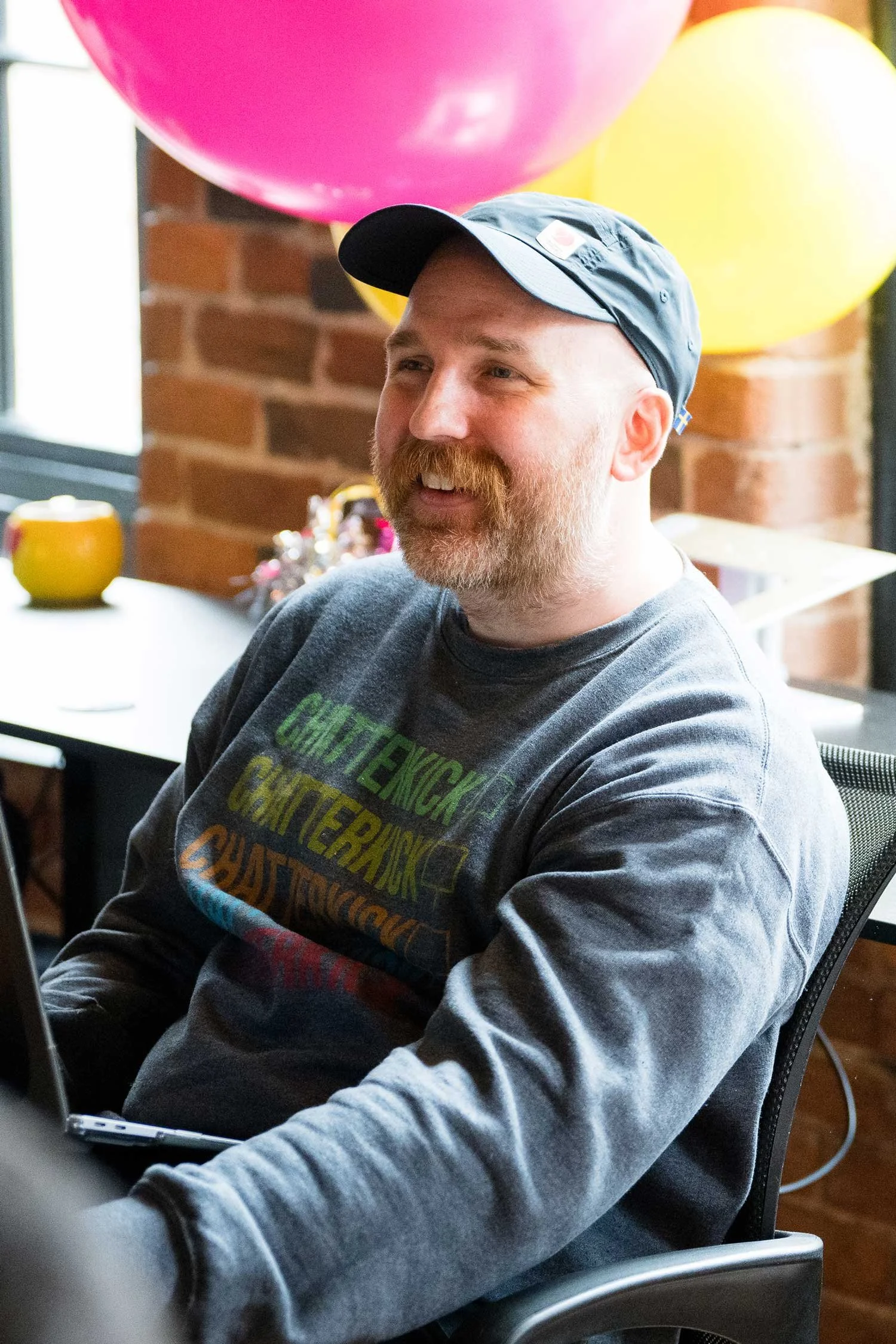 A man with a beard and a cap sitting in a modern office space, smiling, with large balloons in the background and a brick wall.