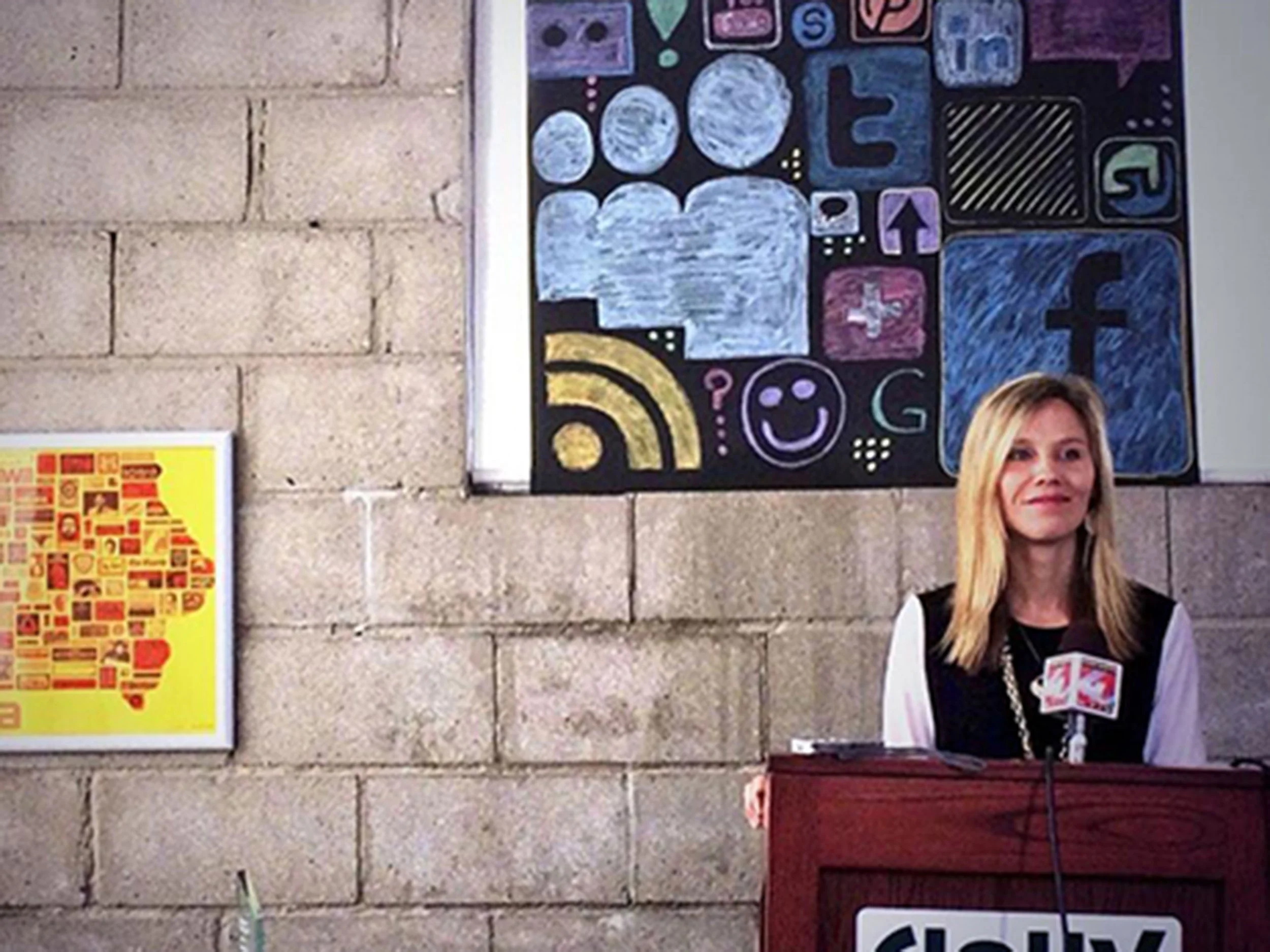 Woman with long blonde hair standing at a podium, smiling, with a chalkboard with social media icons behind her and a yellow framed poster on the brick wall.