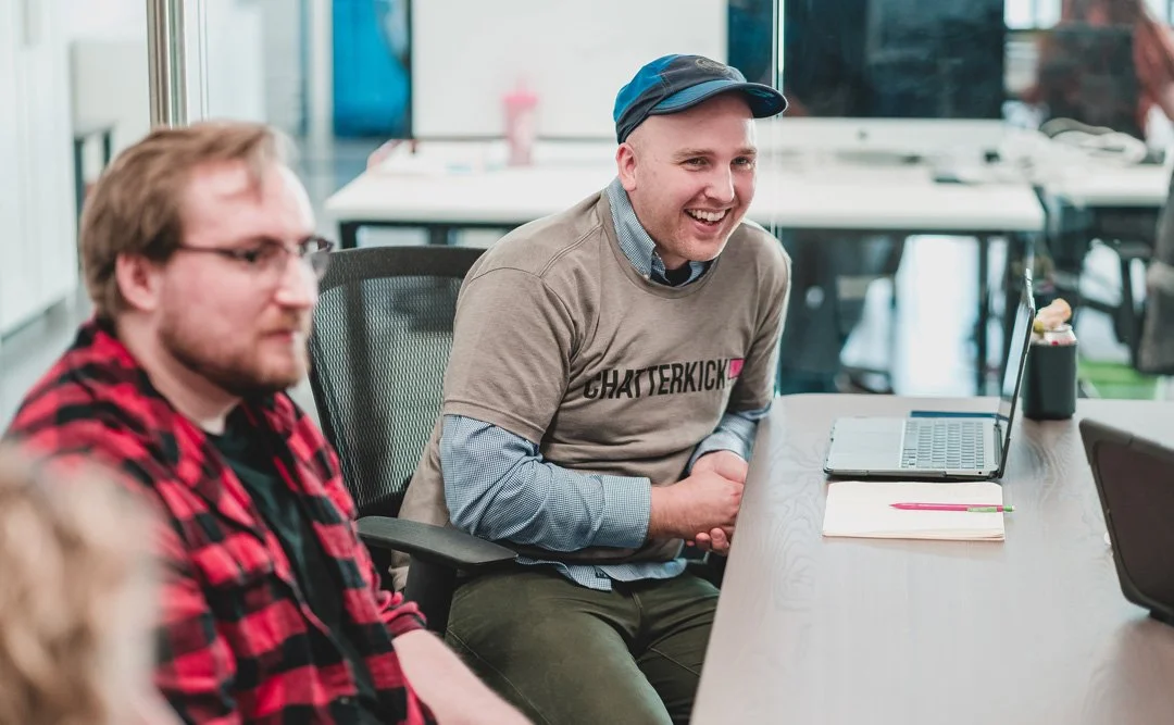 Two men sitting at a conference table in an office, one smiling with a laptop in front of him, the other blurred in the foreground, both engaged in a meeting.