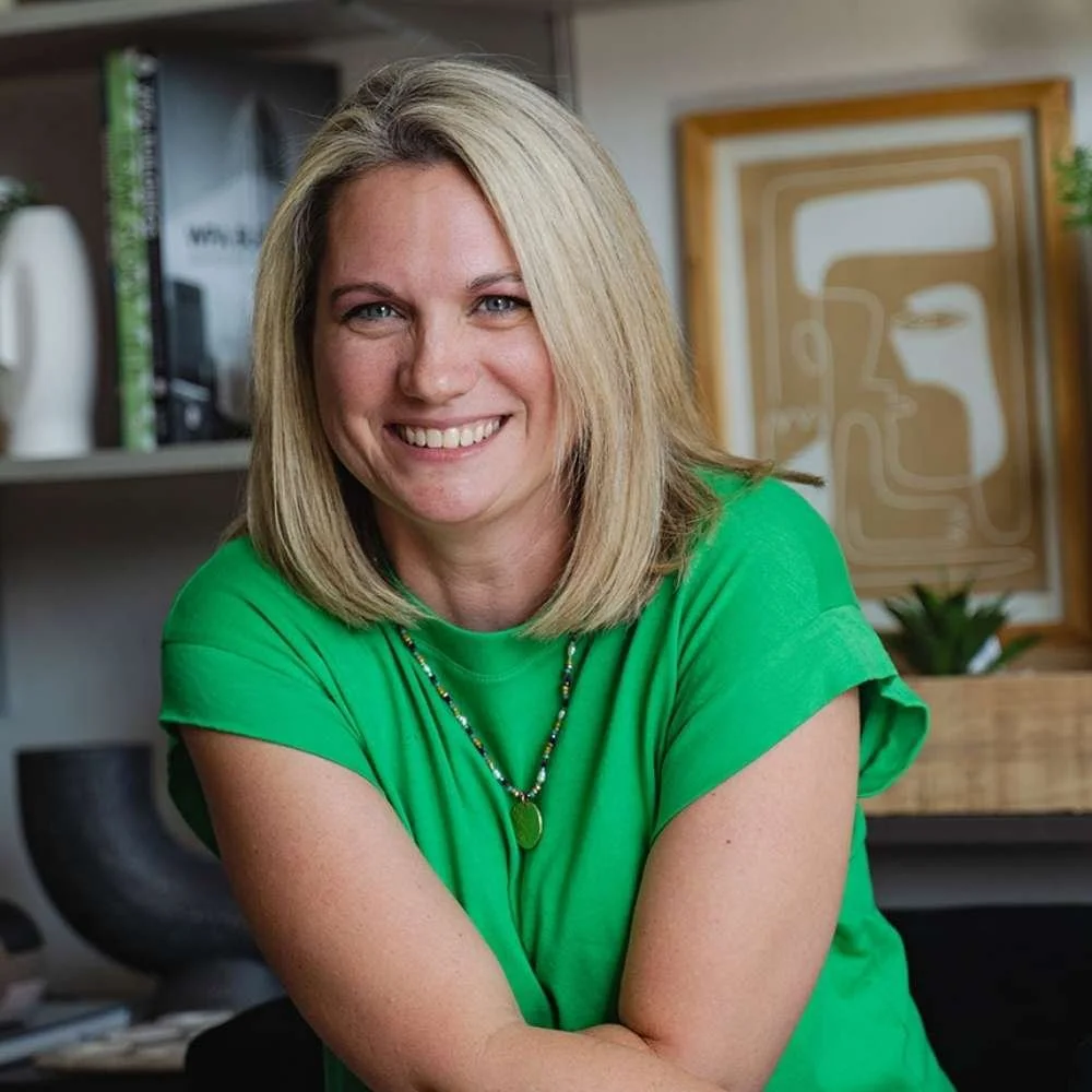A woman with shoulder-length blonde hair, smiling, wearing a green shirt and a beaded necklace, sitting in a room with books, artwork, and decorative items in the background.