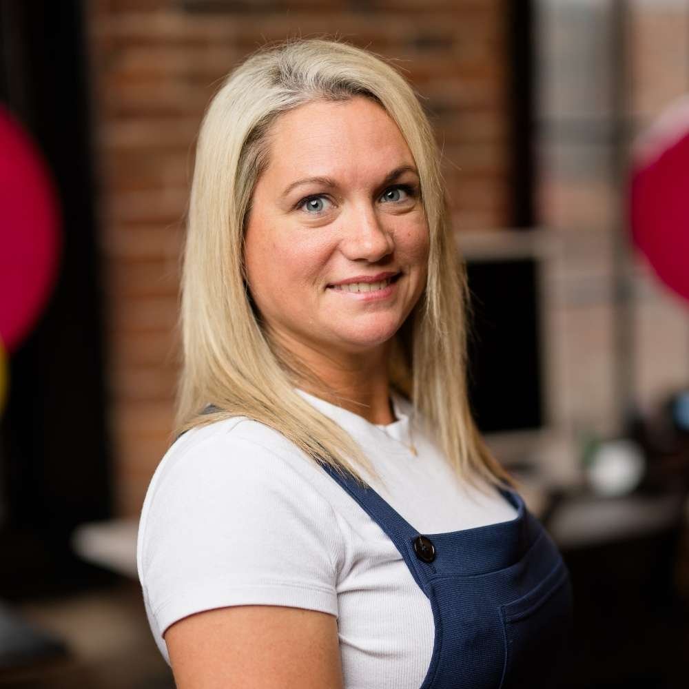 A woman with blonde hair, wearing a white shirt and navy apron, standing inside a room with a brick wall background.