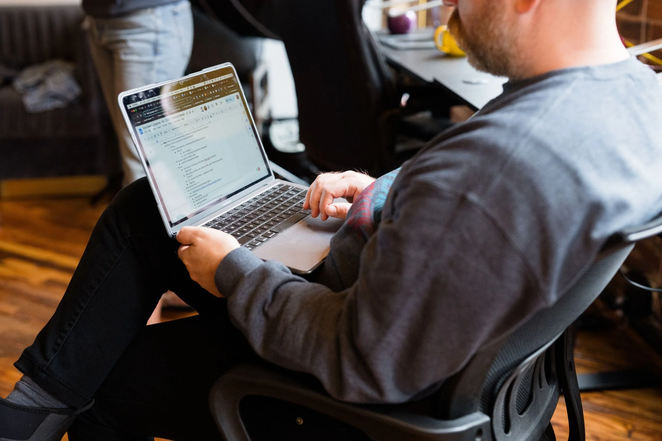 A man sitting in an ergonomic chair working on a laptop in a casual setting, with a wooden floor and other furniture visible.
