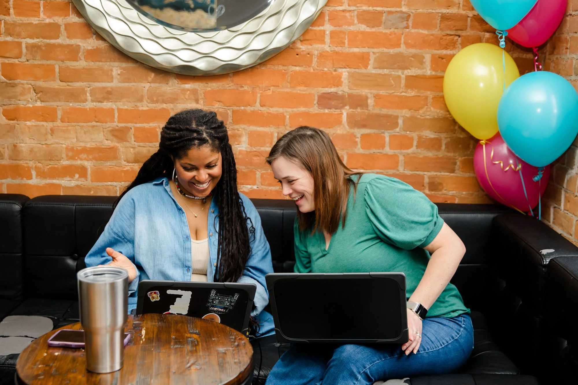 Two women sitting on a black sofa with laptops in front of a brick wall decorated with colorful balloons and a shiny metallic balloon. They are smiling and looking at their laptops.