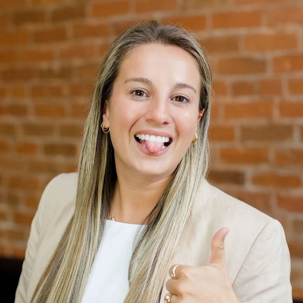 A young woman with long blonde hair smiling and giving a thumbs up gesture in front of a brick wall.