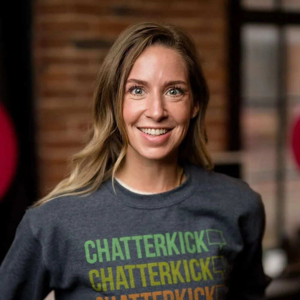 A woman with long light brown hair and blue eyes smiling, wearing a dark gray T-shirt with multiple instances of the word "CHATTERKICK" in green, yellow, and orange text.