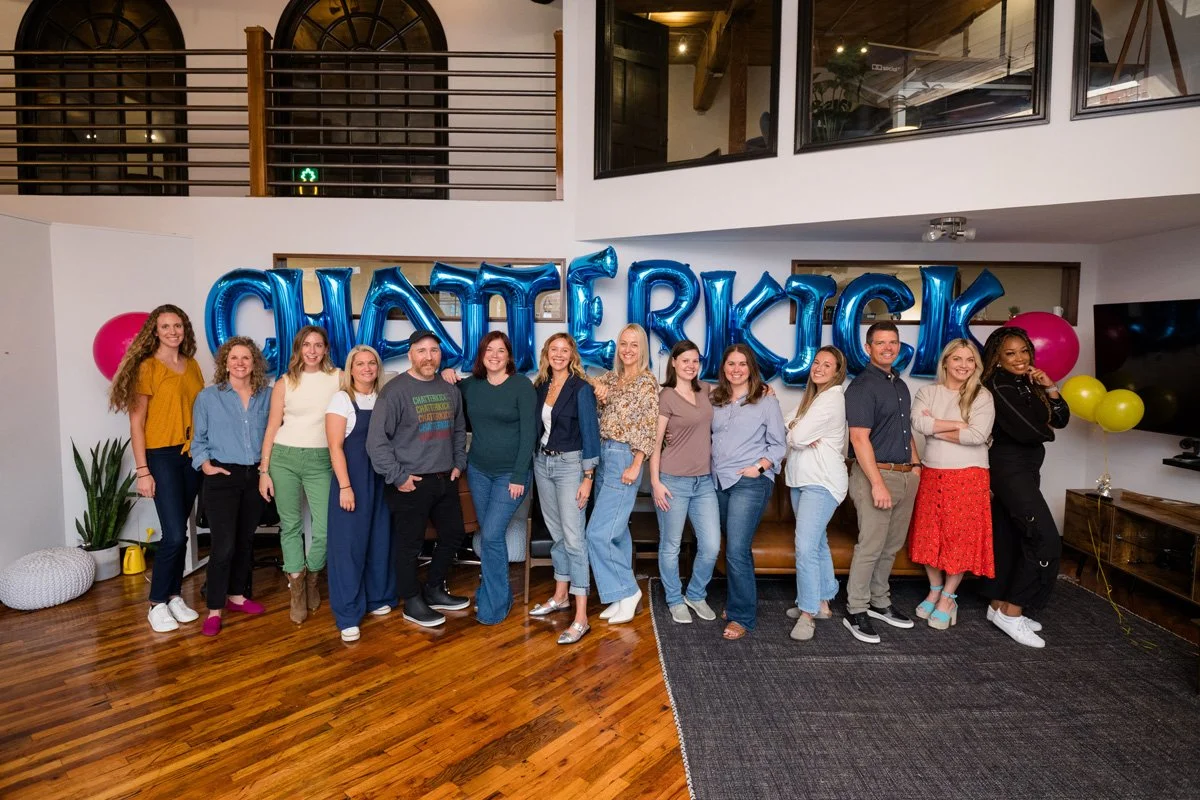 Group of people standing together in a party setting with large blue helium balloons spelling out 'CHATFREAK.' Some are smiling and posing for the photo, with colorful balloons and indoor decor in the background.