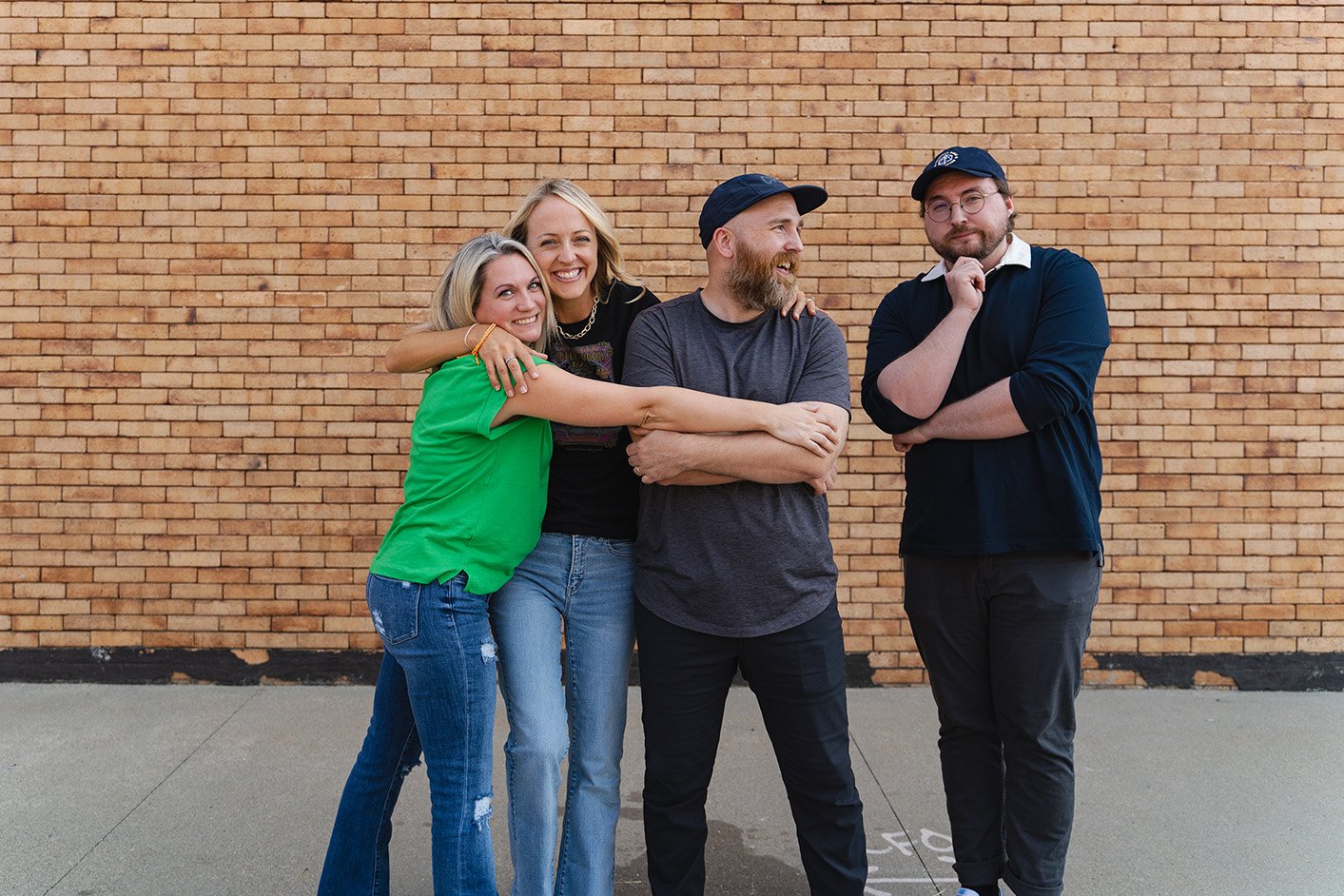 Four friends standing in front of a brick wall, laughing and smiling, with one person hugging another.