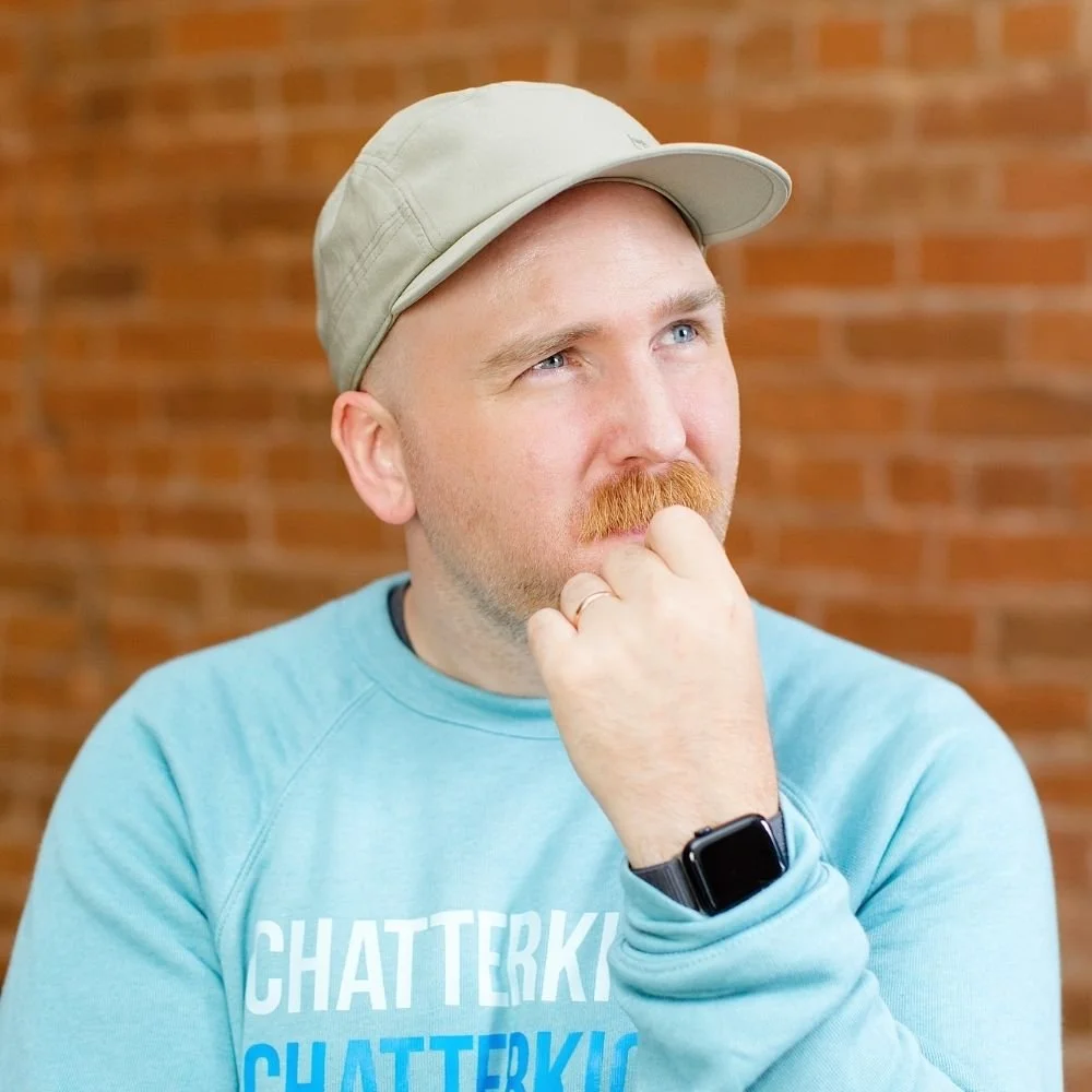 A man with a beige cap, light blue sweatshirt, and a smartwatch, standing against an orange brick wall, appears to be in thoughtful contemplation.
