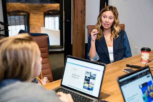 Two women in a meeting room, one speaking and the other listening, laptops on a wooden table, coffee cup on the table.