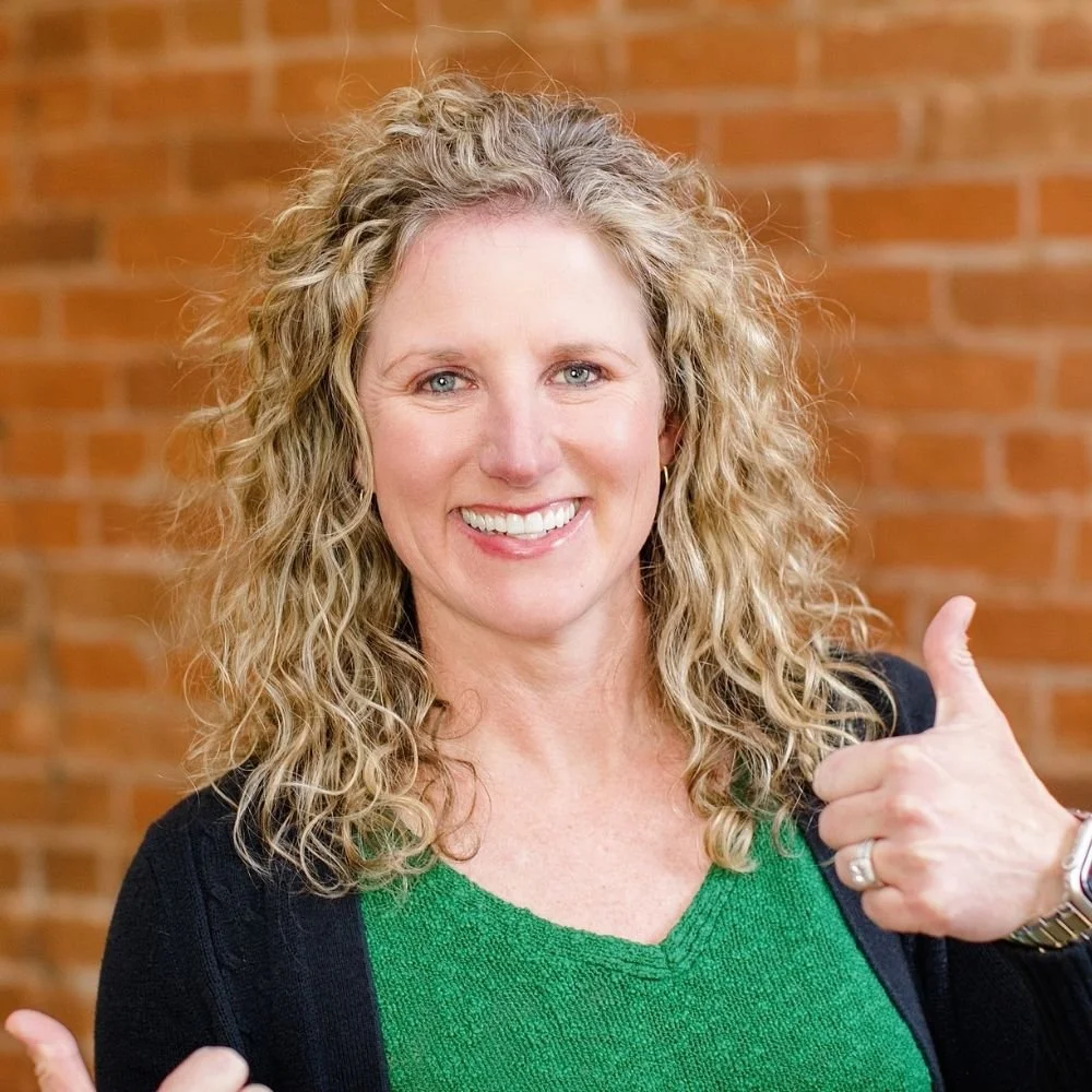 A woman with curly blonde hair smiling and giving a thumbs-up gesture in front of a brick wall.