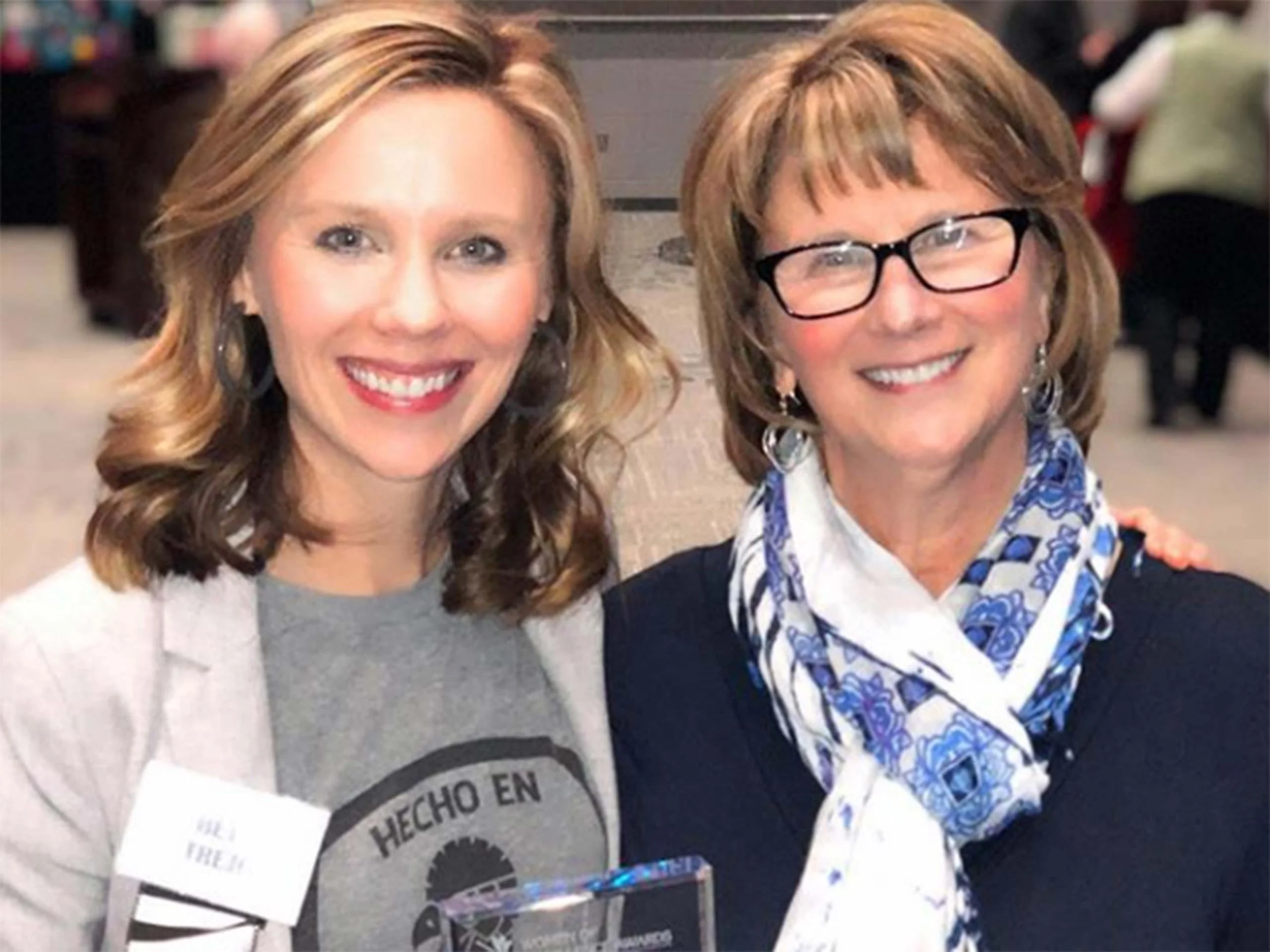 Two smiling women posing for a photograph at an indoor event, one with shoulder-length blonde hair, wearing a light-colored blazer and earrings, and the other with short brown hair, wearing glasses, a dark top, and a blue and white scarf.