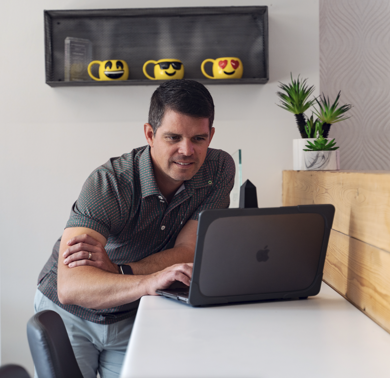 A man looking at a laptop in a modern room with a shelf holding emoji mugs and a decorative plant.